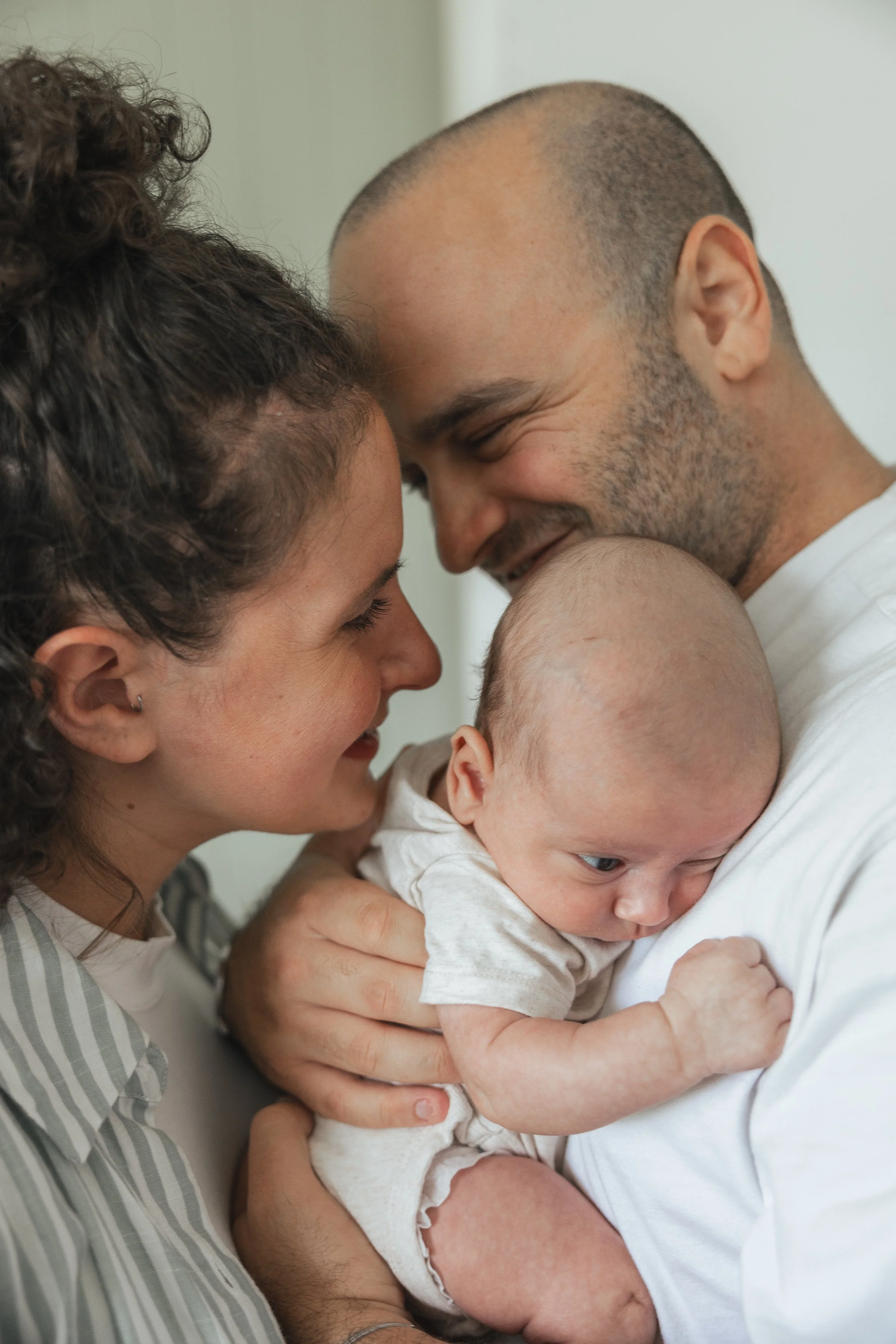 Close-up of a couple holding their baby and smiling at each other, with their foreheads touching.
