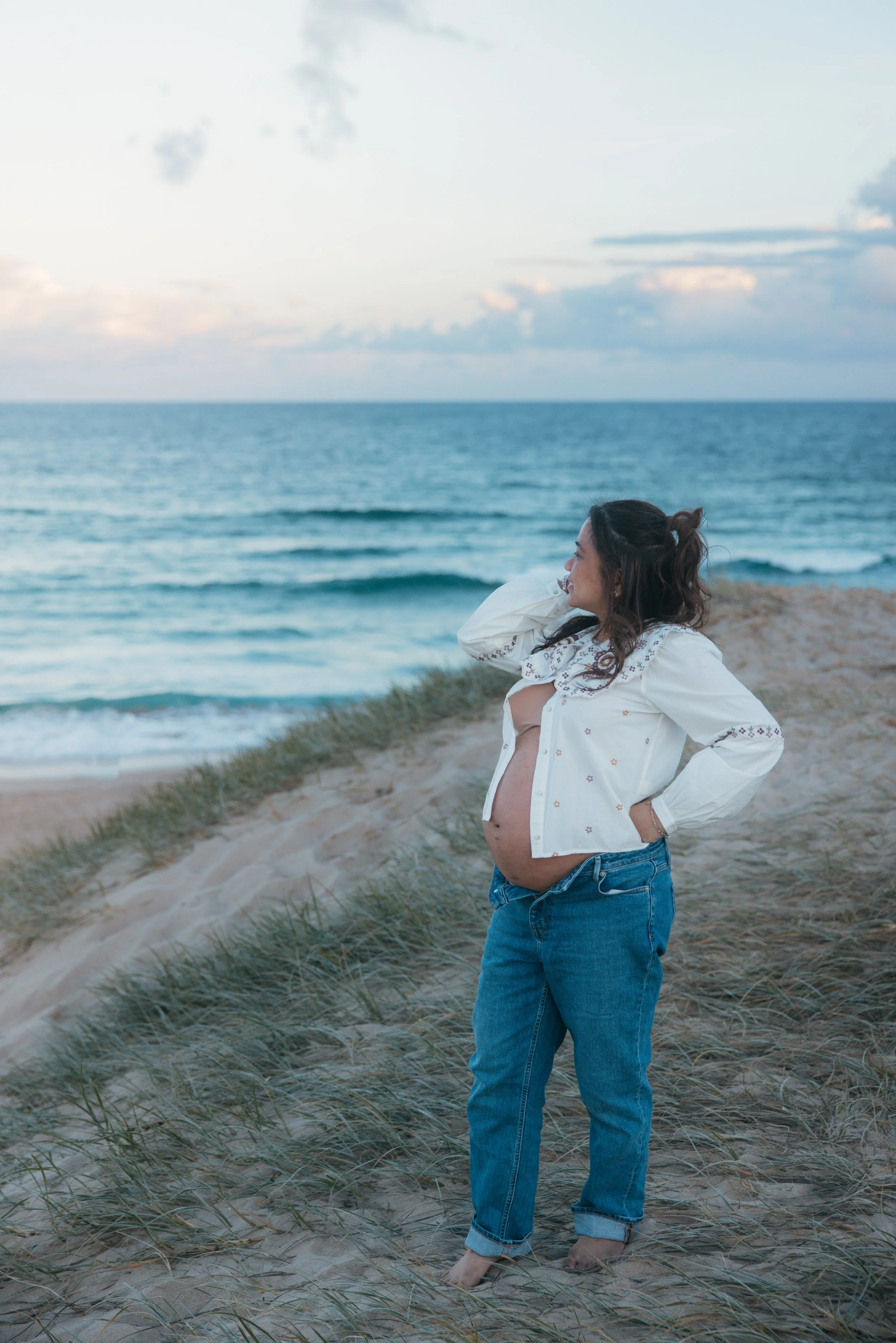 A pregnant woman stands on a sandy beach with green grass, looking out at the ocean with waves and a cloudy sky.