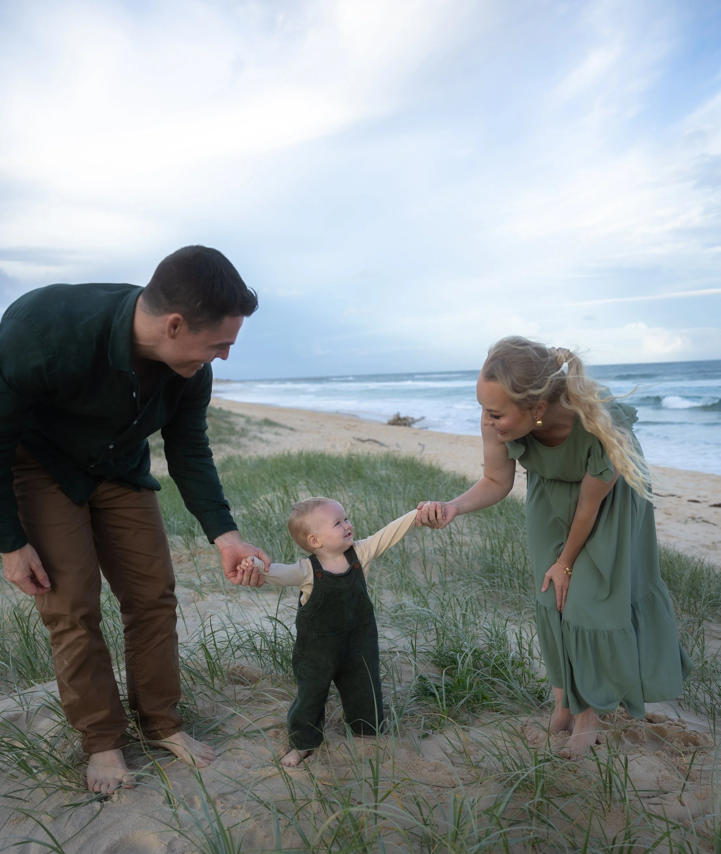 A family of three at the beach holding hands, with the child in the middle and the parents smiling at each other.