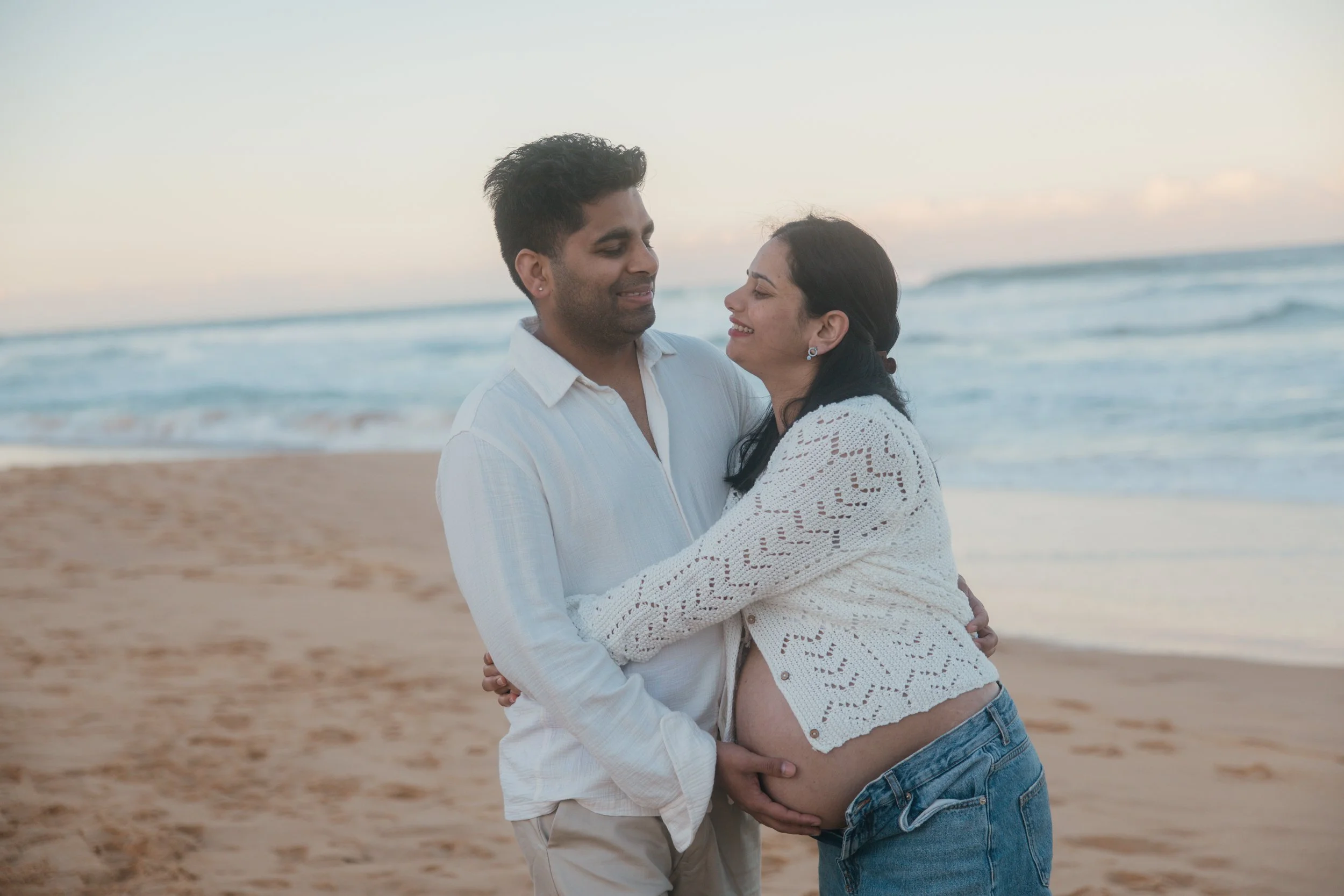 A smiling pregnant woman in a white, crochet top and jeans embraces a man on a beach during sunset, both wearing white shirts.