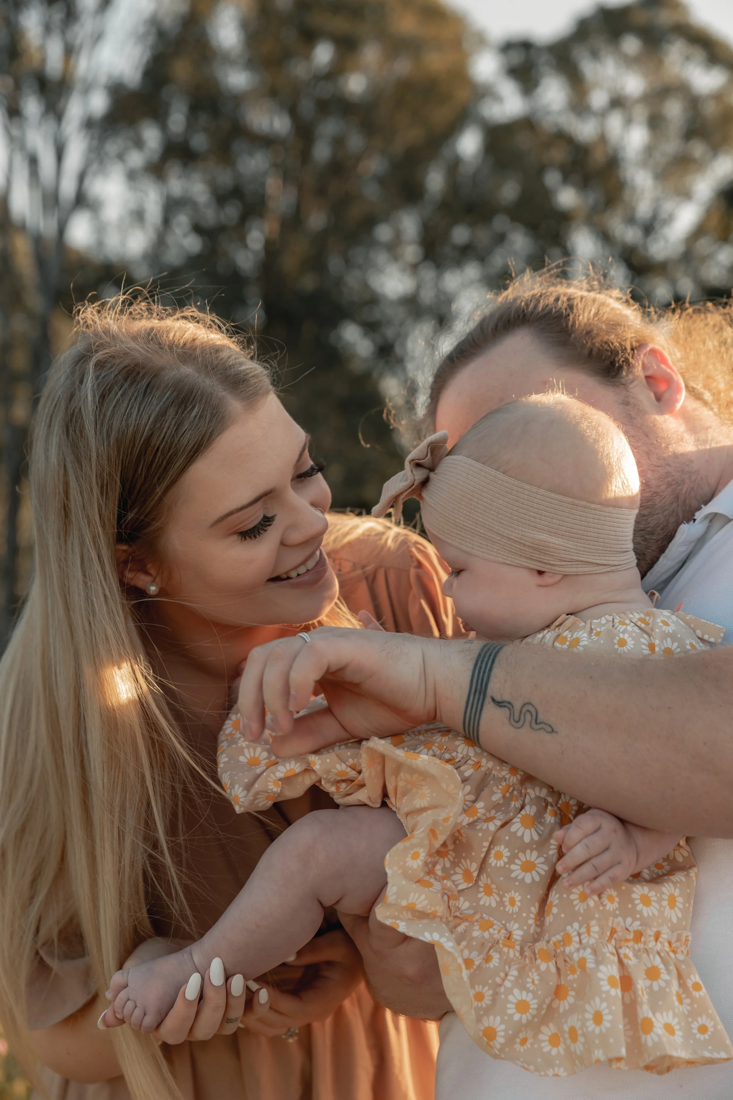 A smiling woman with long blonde hair holding a baby girl dressed in a daisy-patterned dress, with another person kissing her on the cheek, outdoors during golden hour.