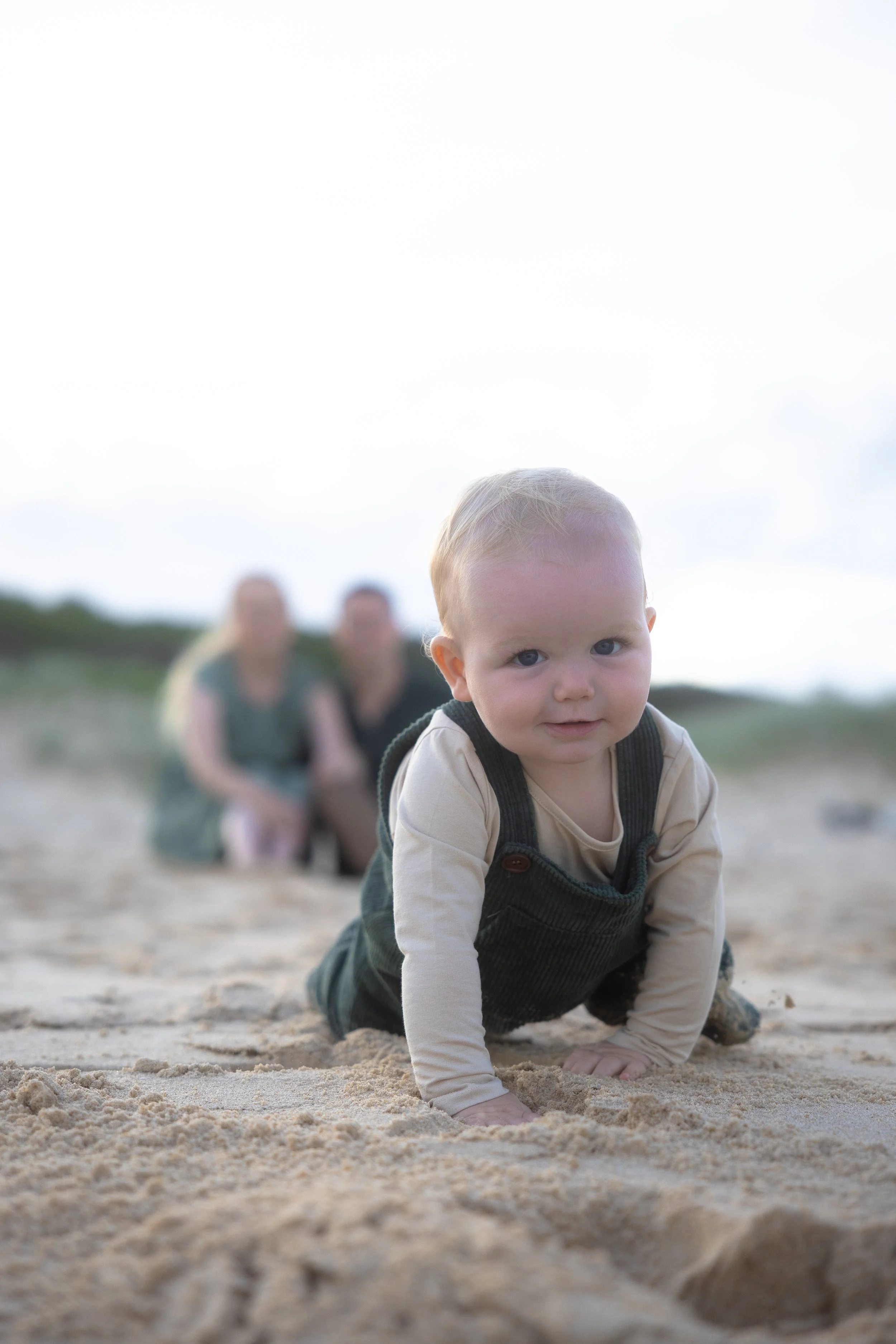 A young child crawling on sandy ground with two women sitting in the background, outdoors under a cloudy sky.