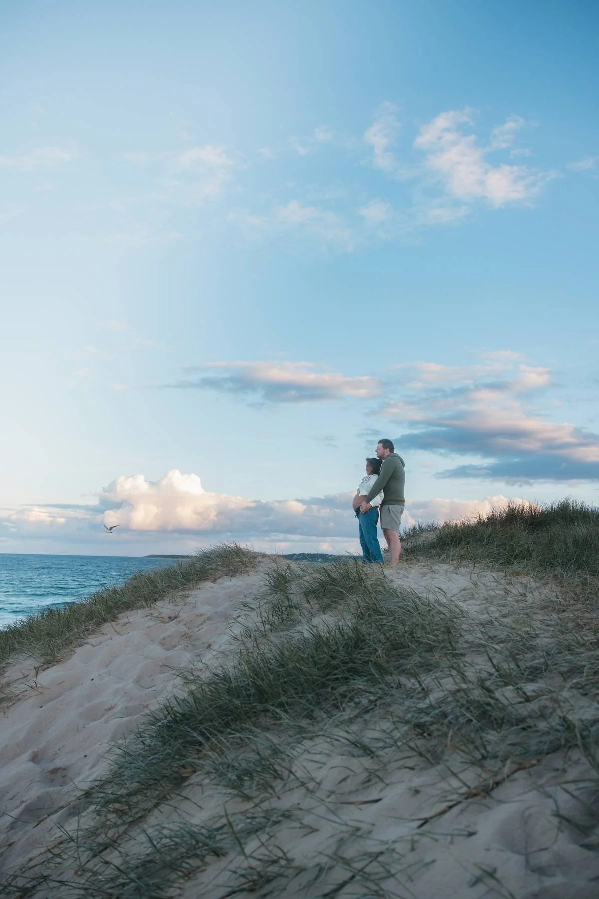 A couple standing on sand dunes by the ocean, watching the sky at sunset.