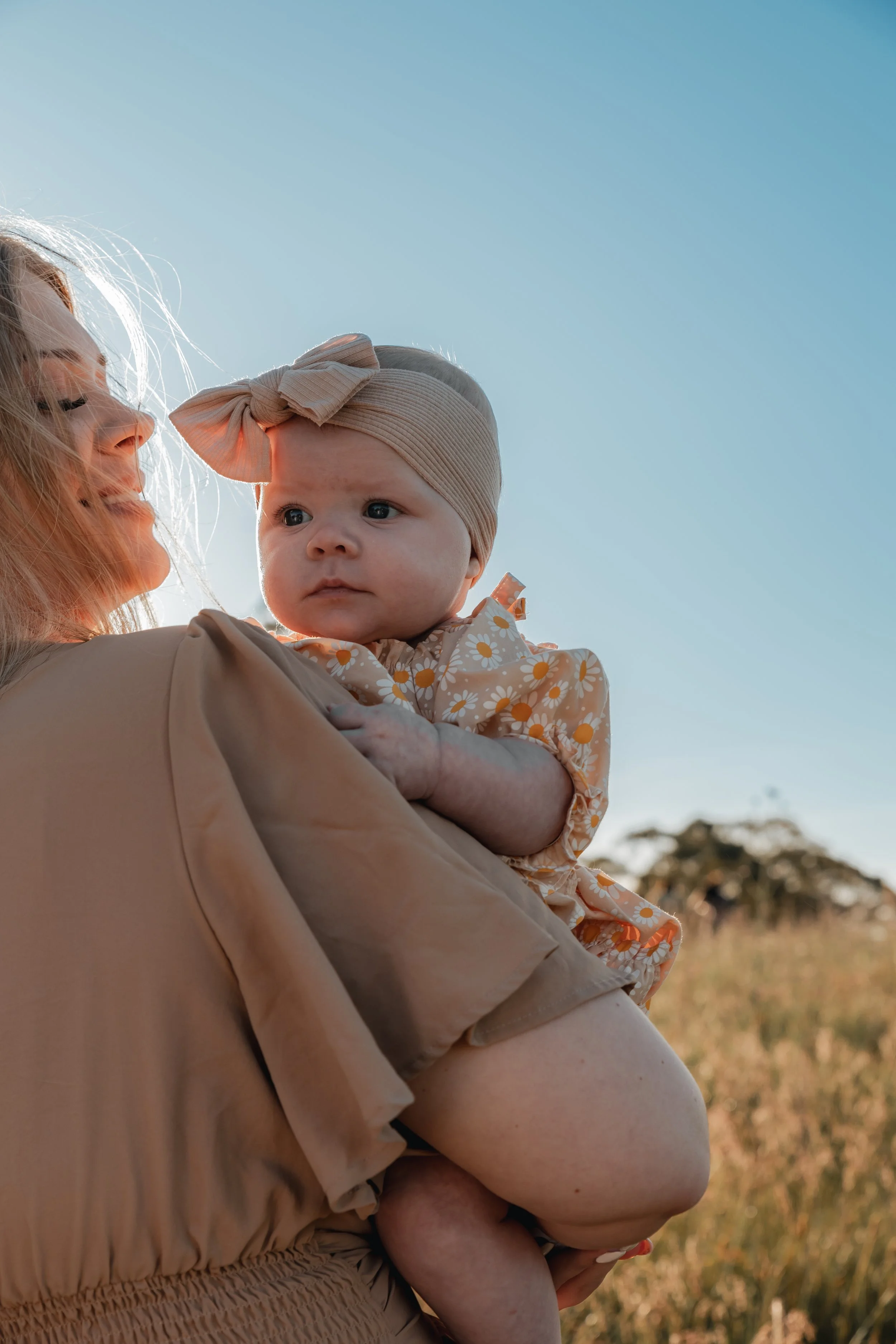 A woman holding a baby outdoors during sunset, with grassy fields and trees in the background.
