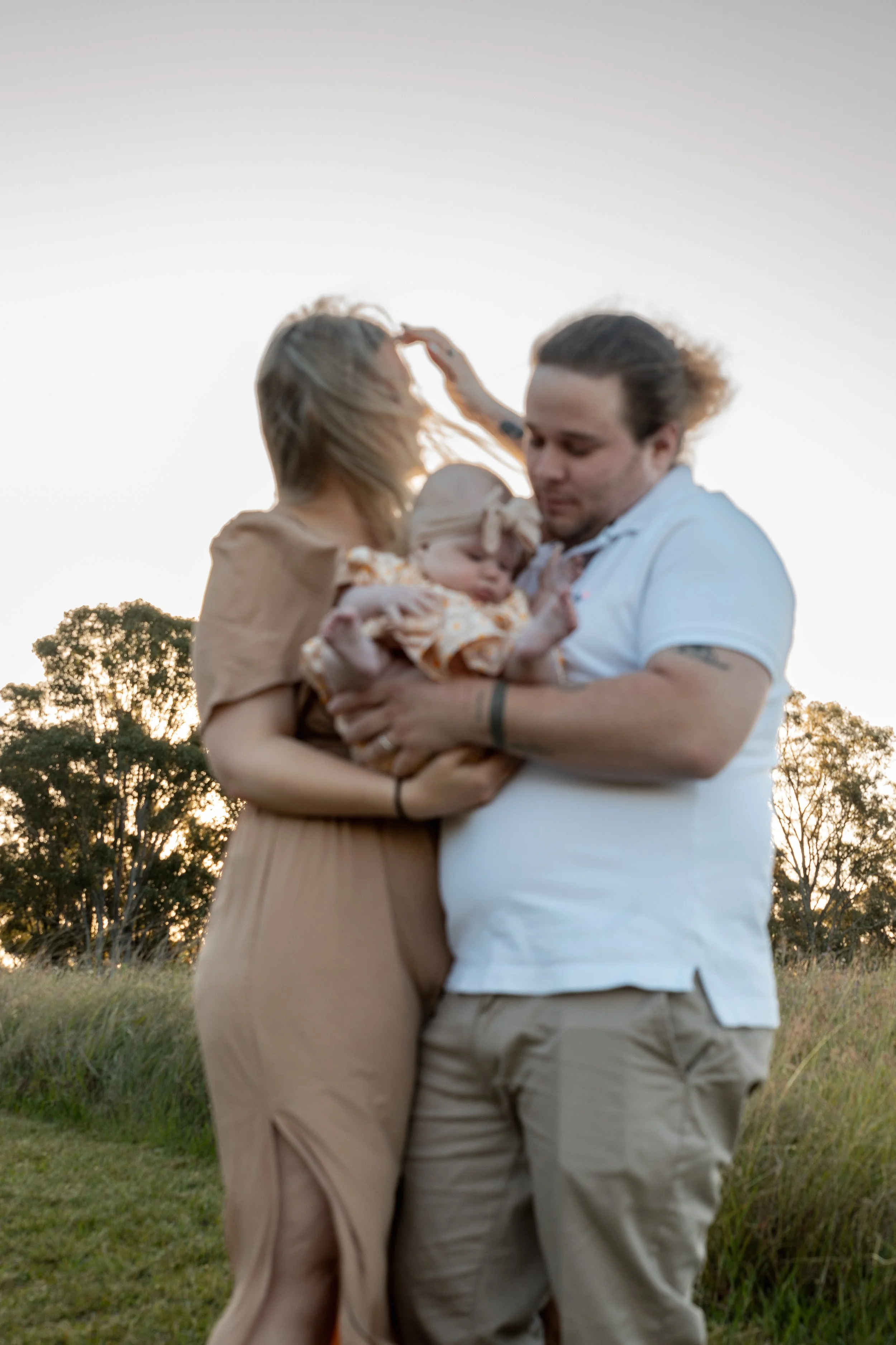 A family outdoors at sunset, with a woman, man, and baby girl. The woman has blonde hair and is wearing a beige dress, holding the baby. The man has dark hair and is wearing a white shirt and beige pants, holding the woman and baby. They are touching
