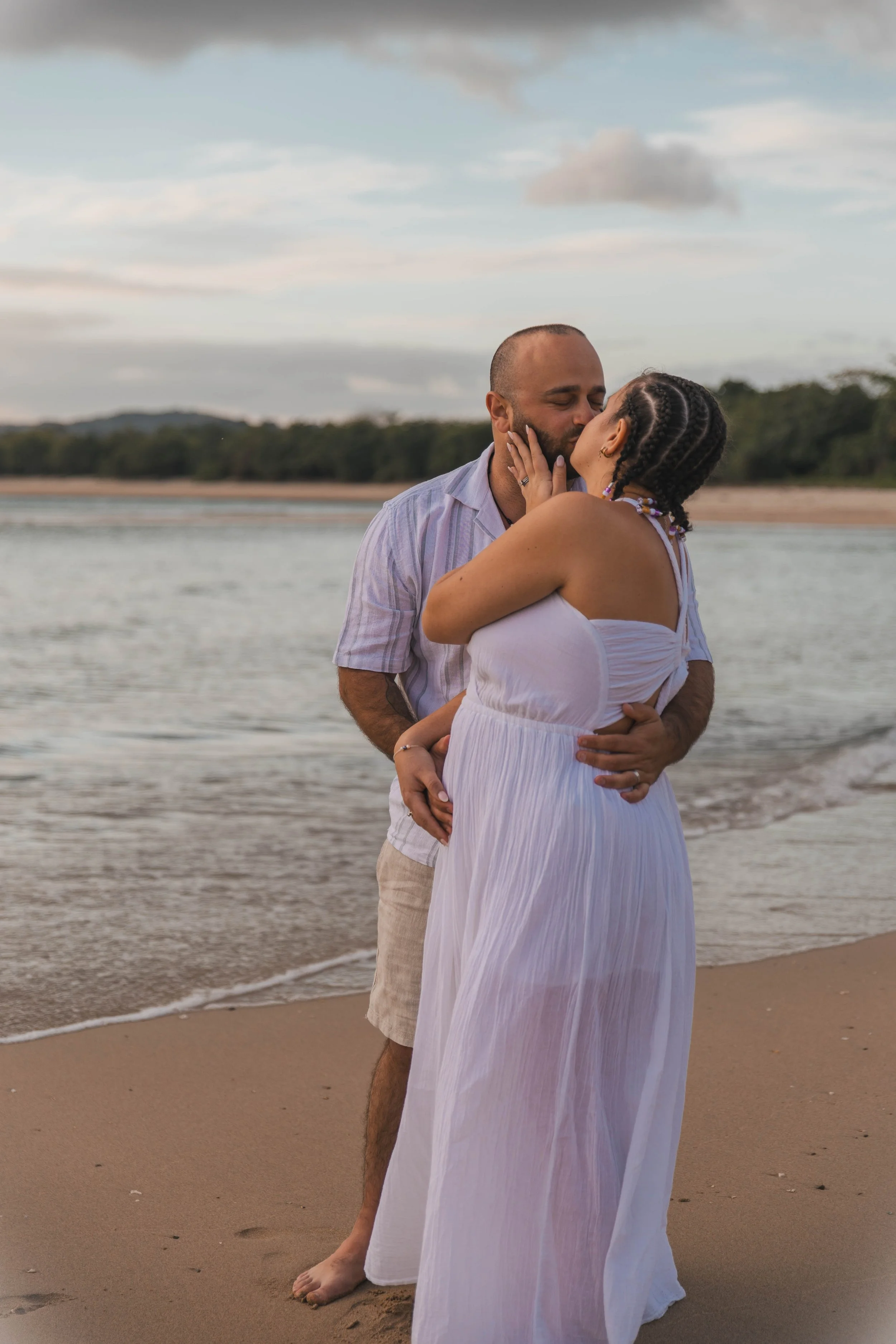 A couple kissing on the beach during sunset, with water and trees in the background.