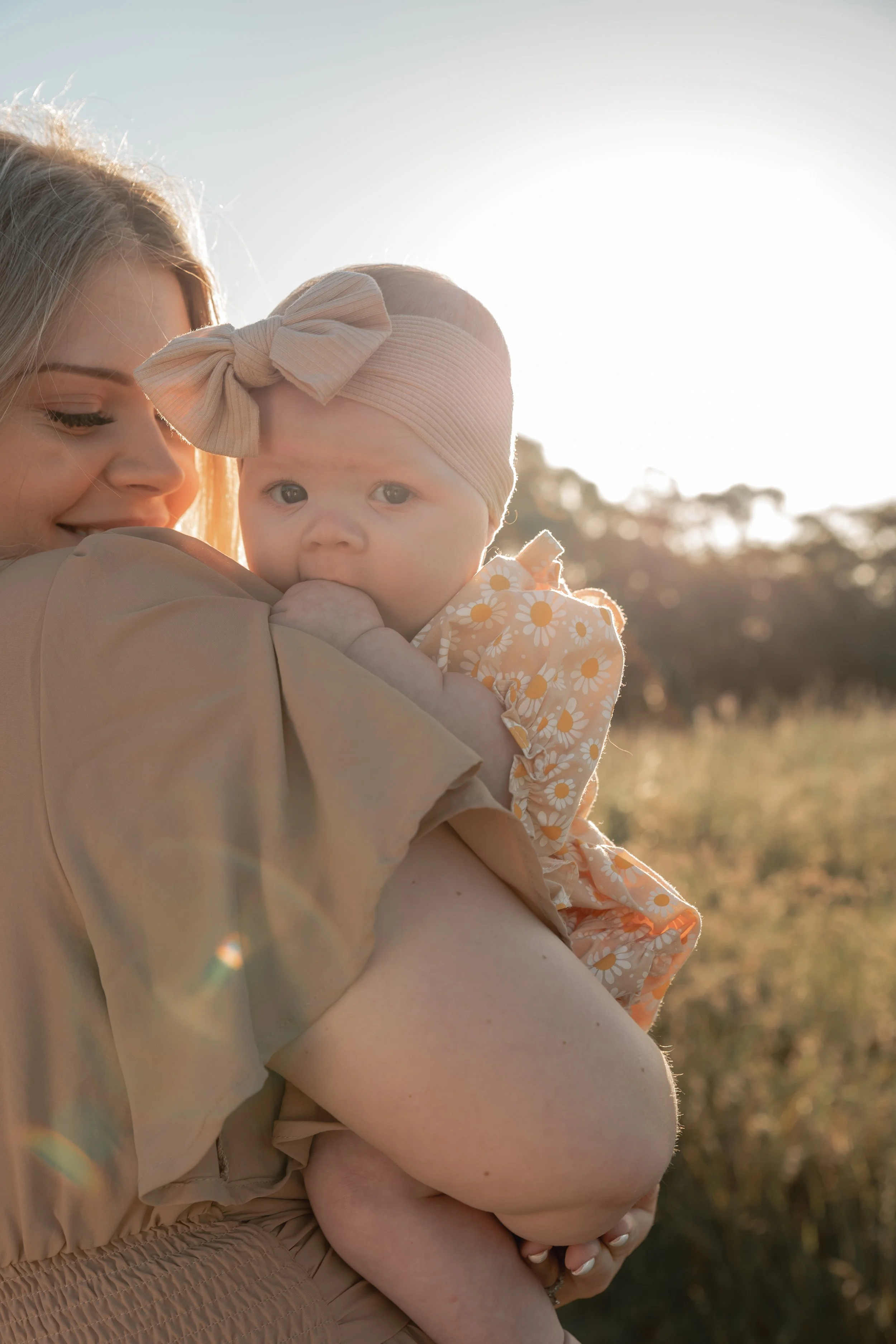 A woman holding a baby girl outdoors during sunset, with the sun shining behind them and a backdrop of trees and grass.