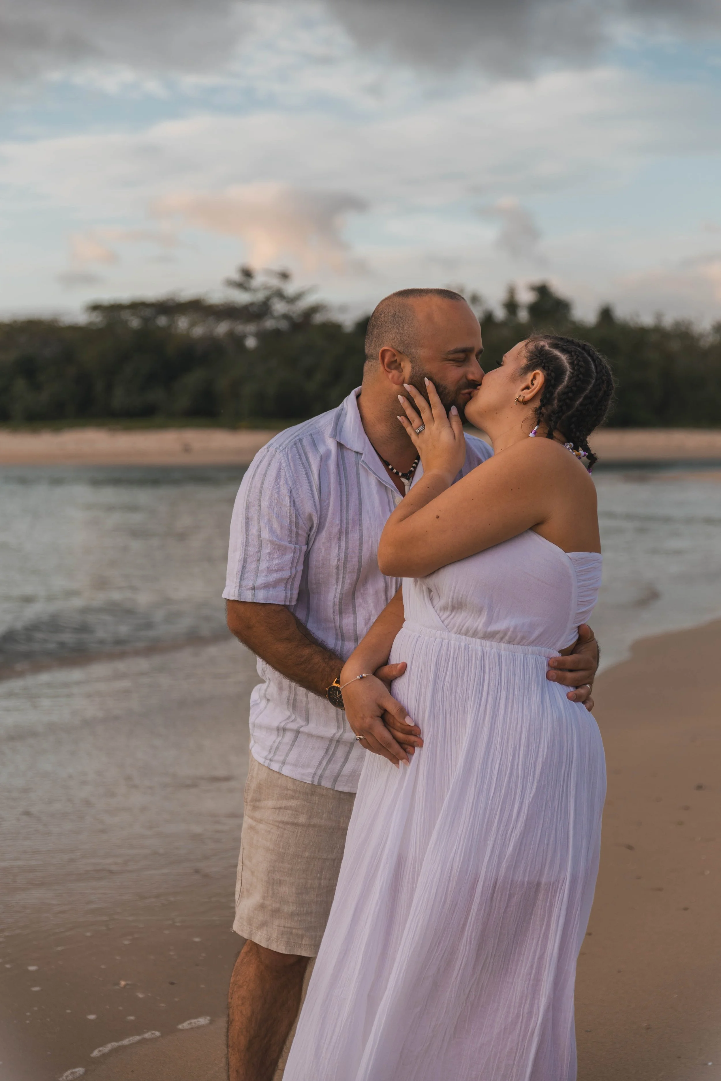 A couple kissing on the beach during sunset, with water, sand, and trees in the background.