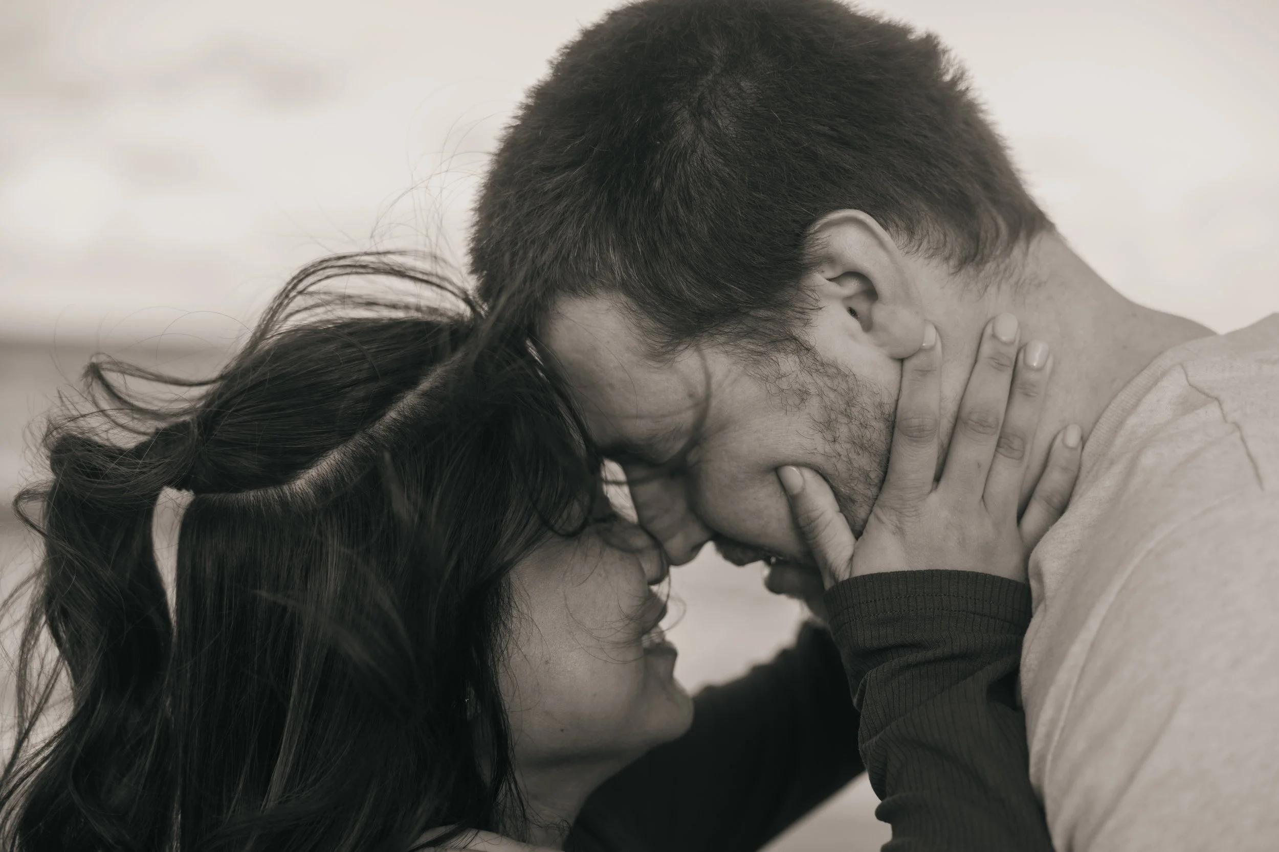 A black-and-white photo of a couple with their foreheads and noses touching, eyes closed, holding each other closely, conveying intimacy and affection.