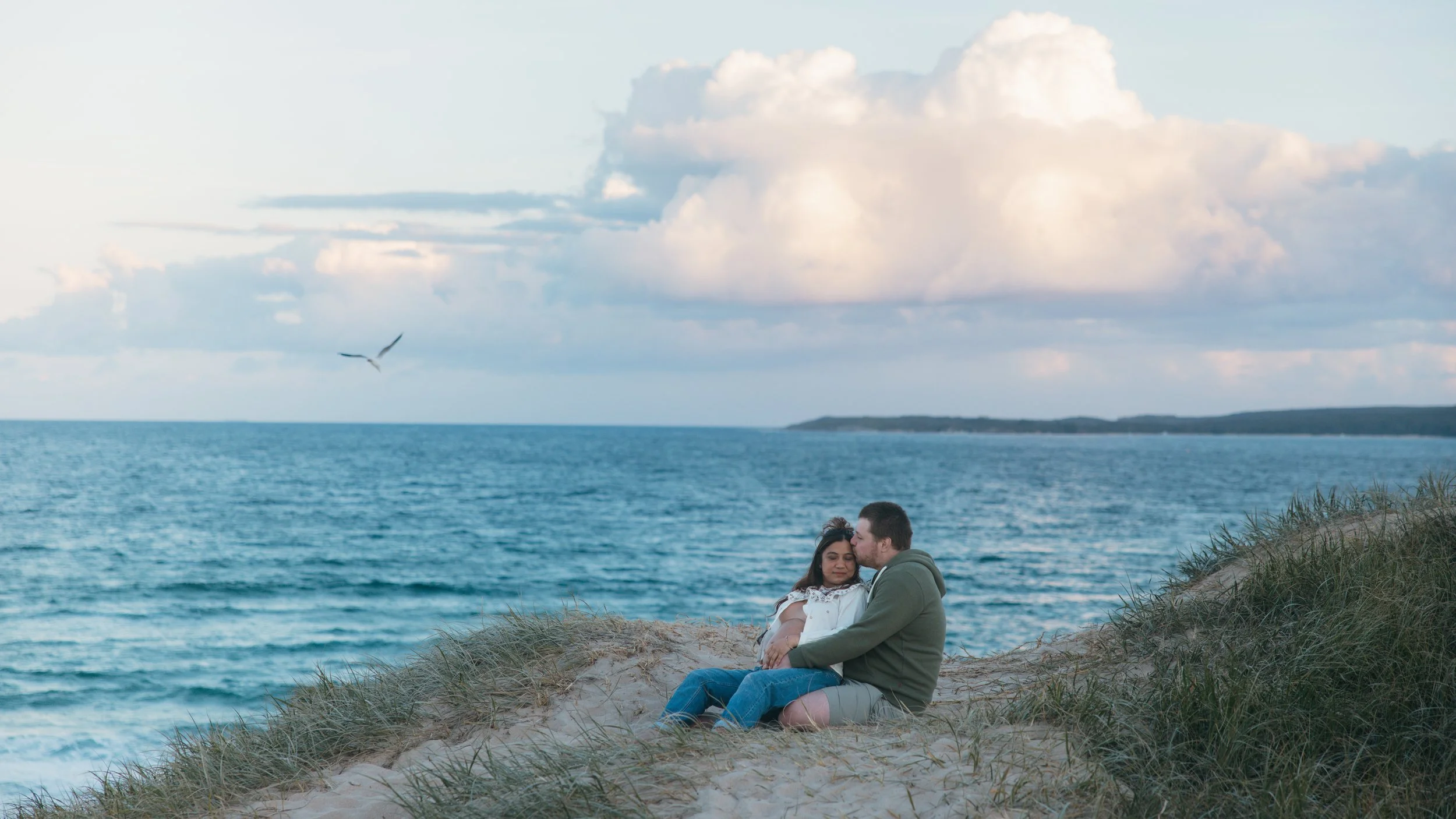 A couple sitting on a grassy sand dune near the ocean, with the man kissing the woman's forehead, under a partly cloudy sky.