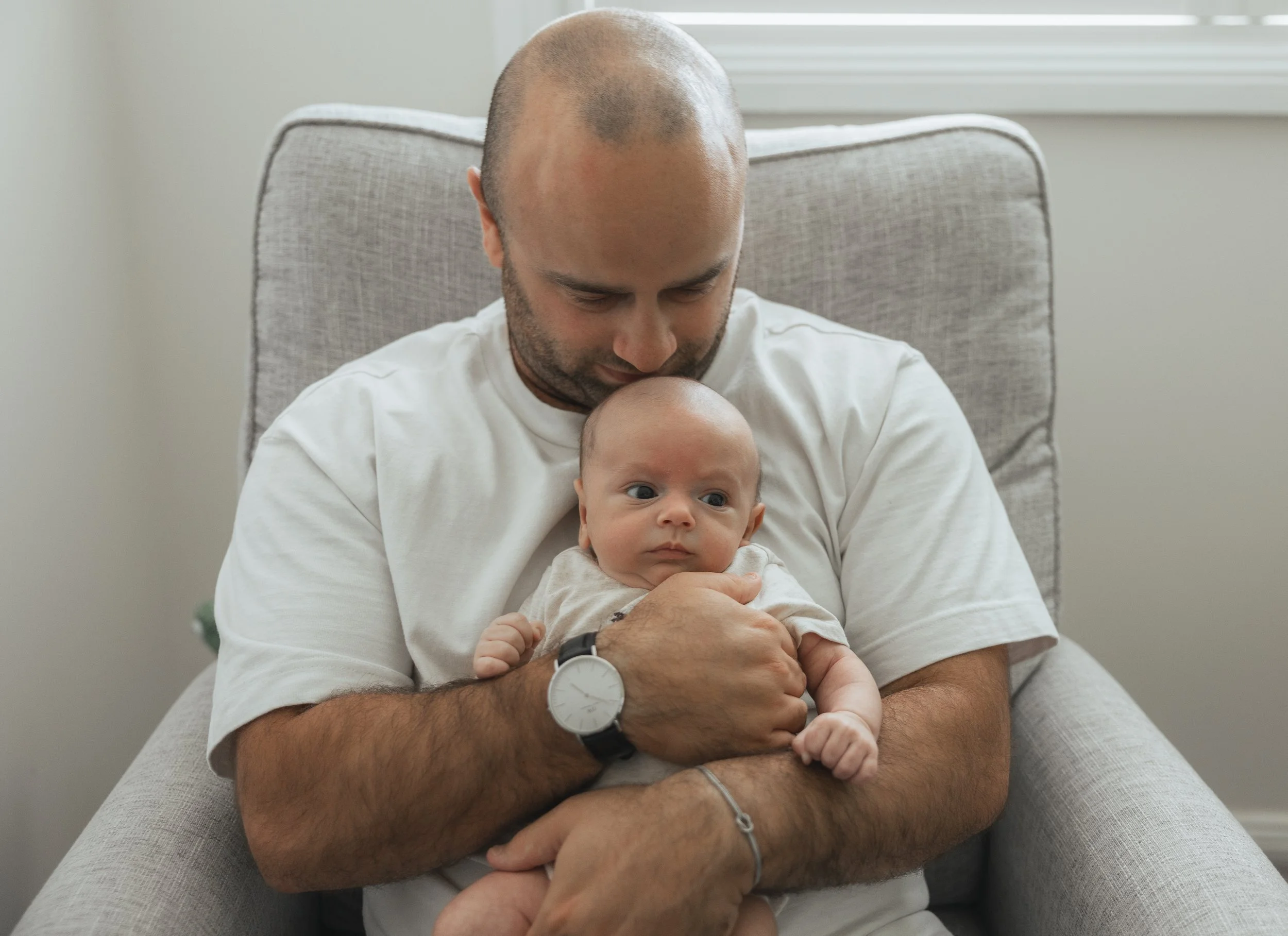 A man holding a baby while sitting in a gray armchair in a well-lit room.