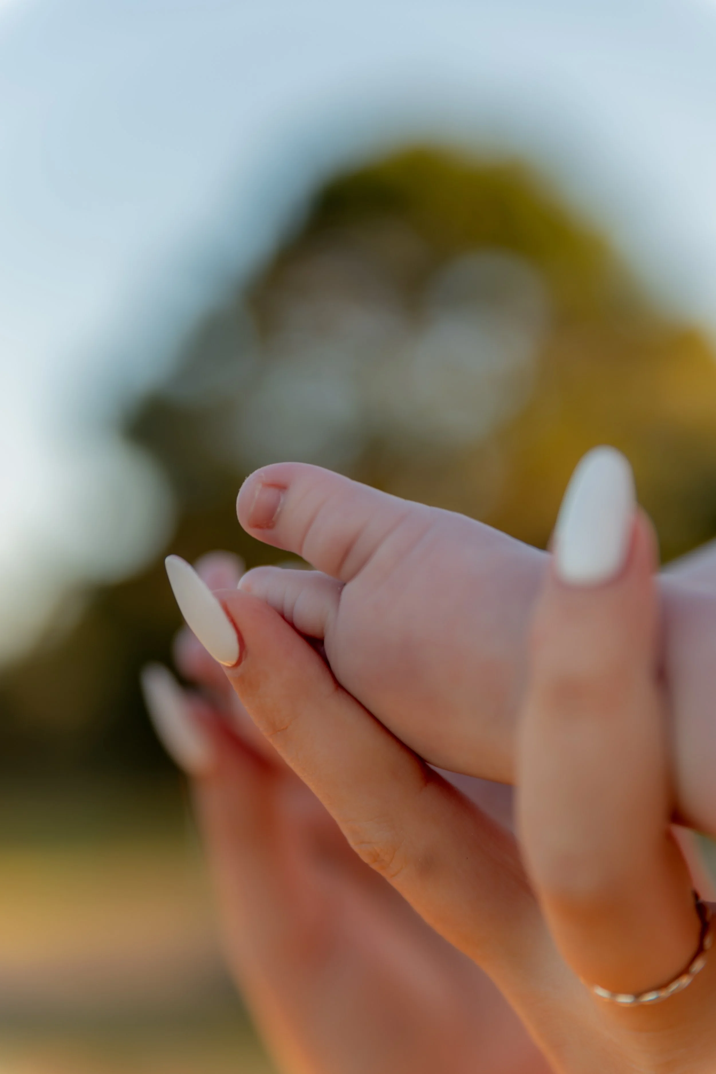 Close-up of a woman’s hand with white manicured nails, lightly holding her own hand, outdoors with blurred trees in the background.