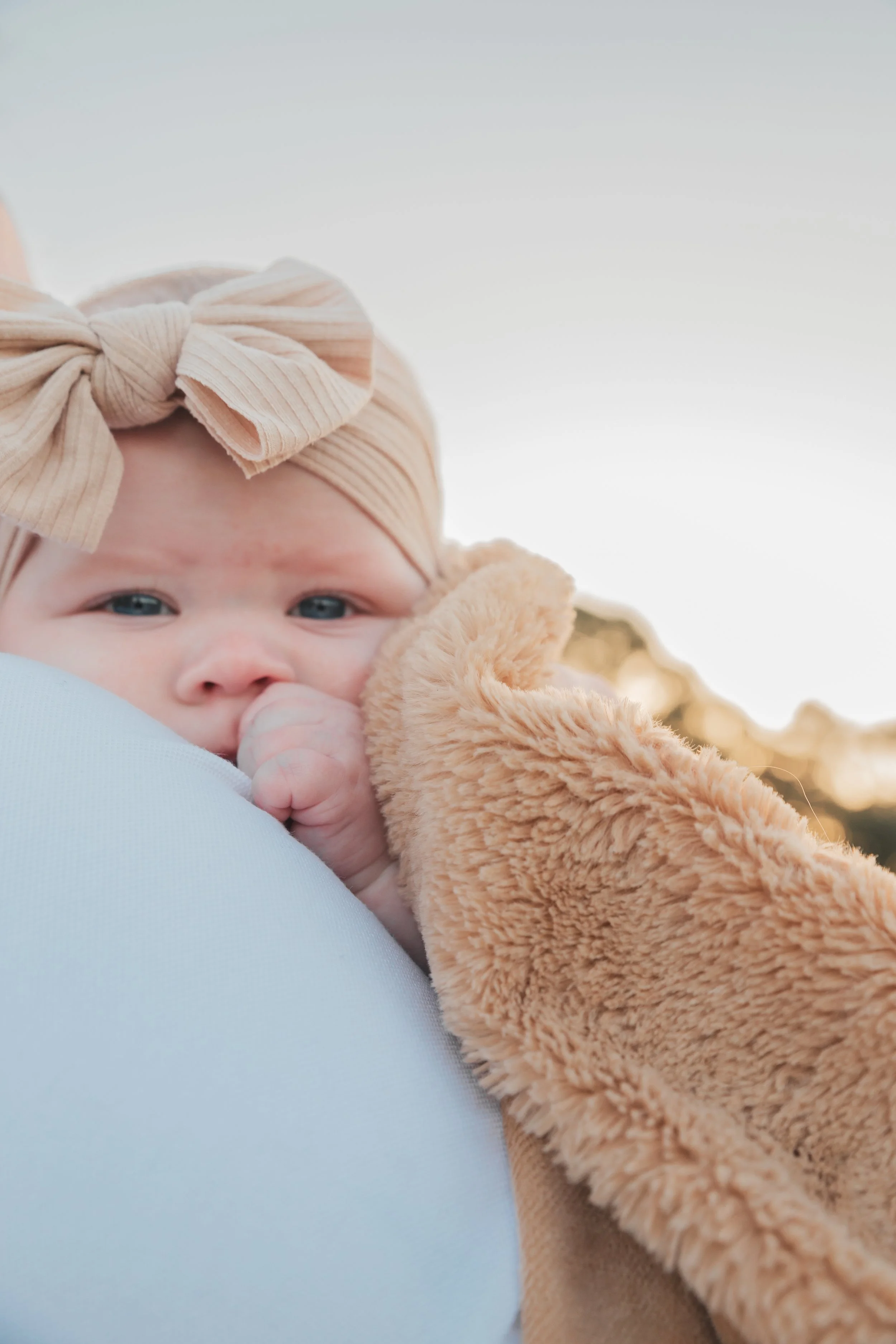 Baby with a beige headband and bow, wrapped in a plush tan blanket, resting on someone’s shoulder outdoors during sunset.