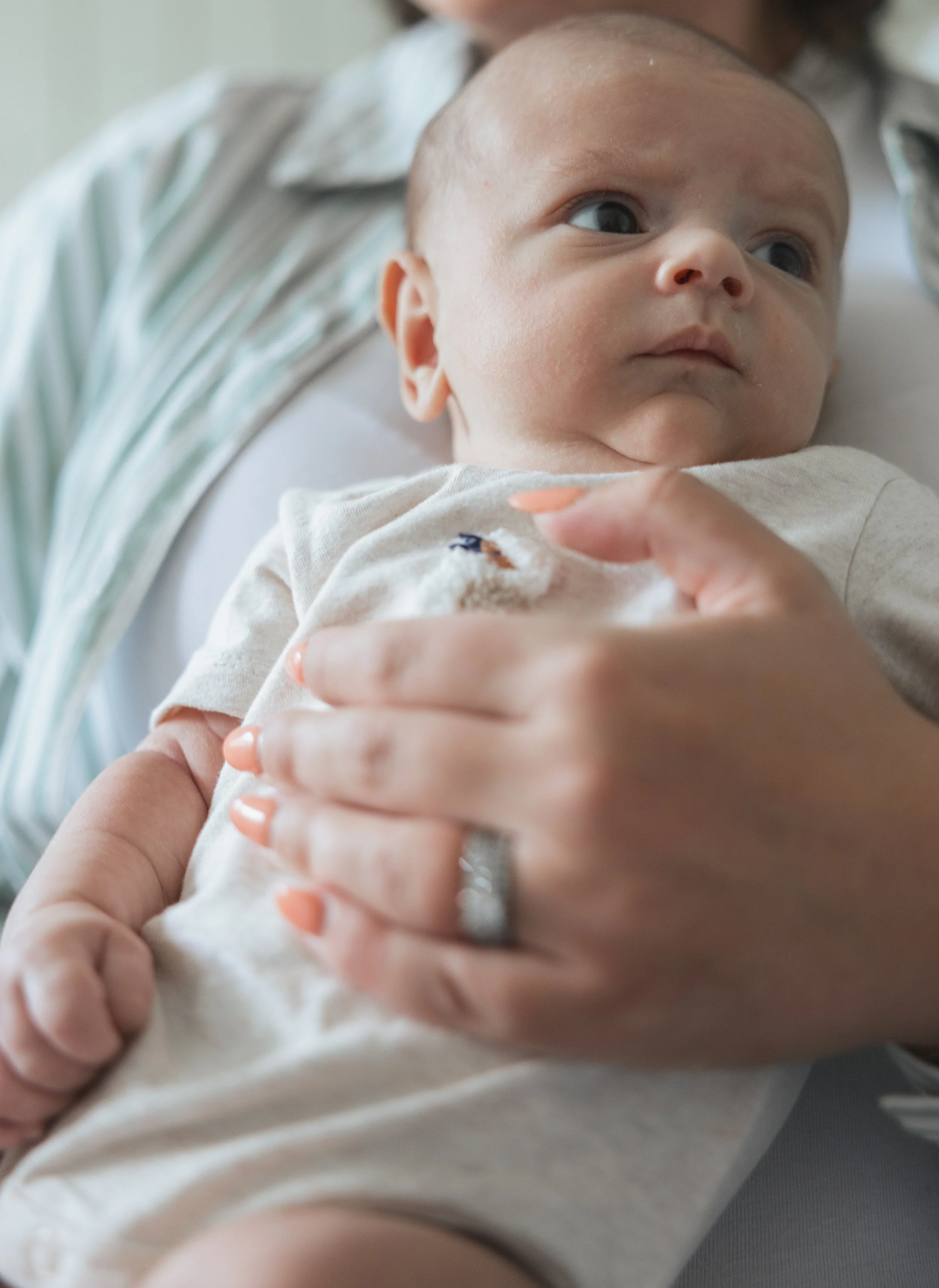 A baby being held by a woman, looking to the side with wide eyes, wearing a light-colored outfit.