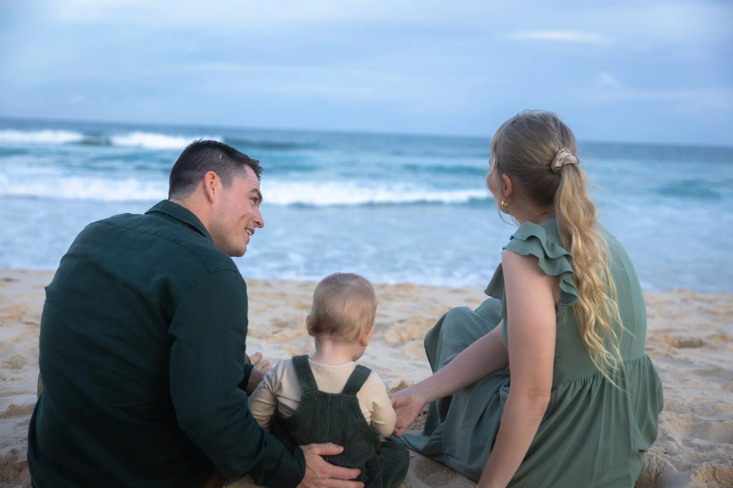 A family of three, including a man, a woman, and a young child, sitting on the beach facing the ocean, enjoying a moment together.