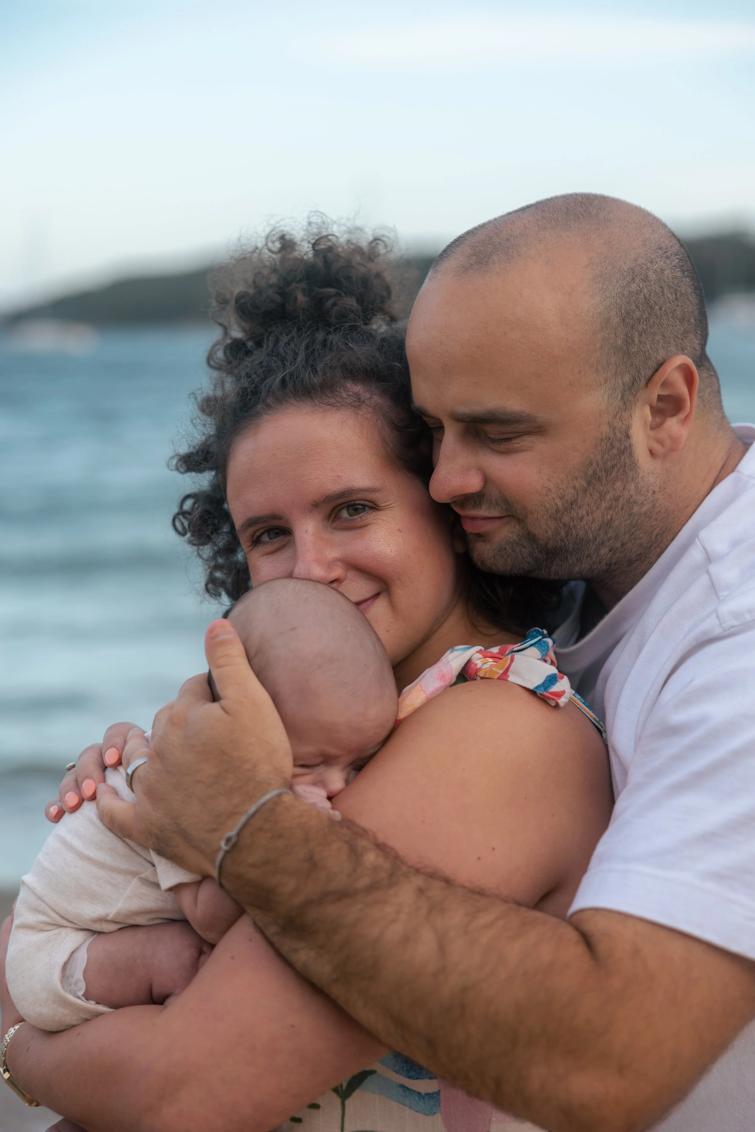 A family with a mother, father, and a baby, embracing and sharing a tender moment on the beach with water and distant land in the background.