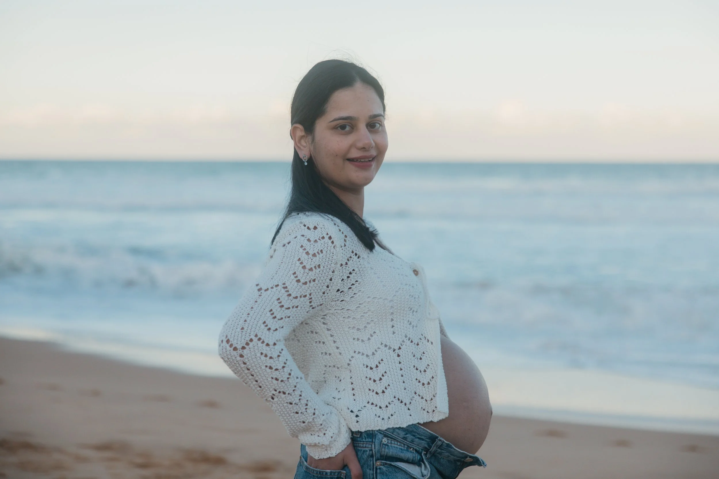 Young pregnant woman standing on a beach with ocean in the background, wearing a white crochet top and jeans.