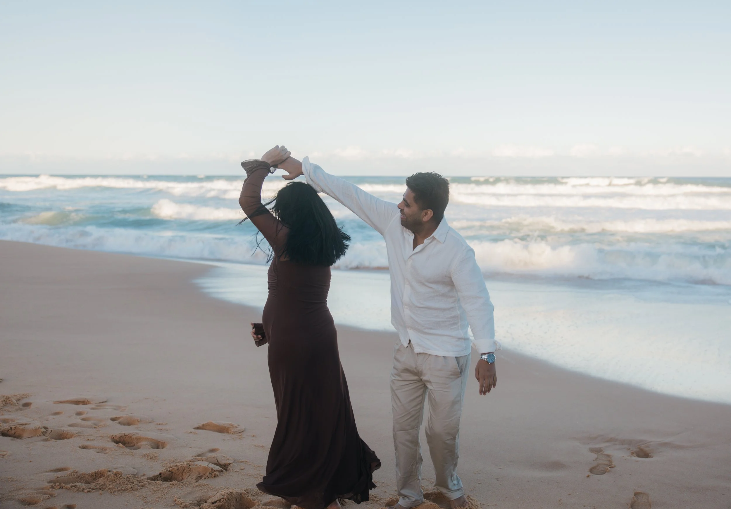 A couple dancing on the beach near the ocean, with waves in the background and footprints in the sand.