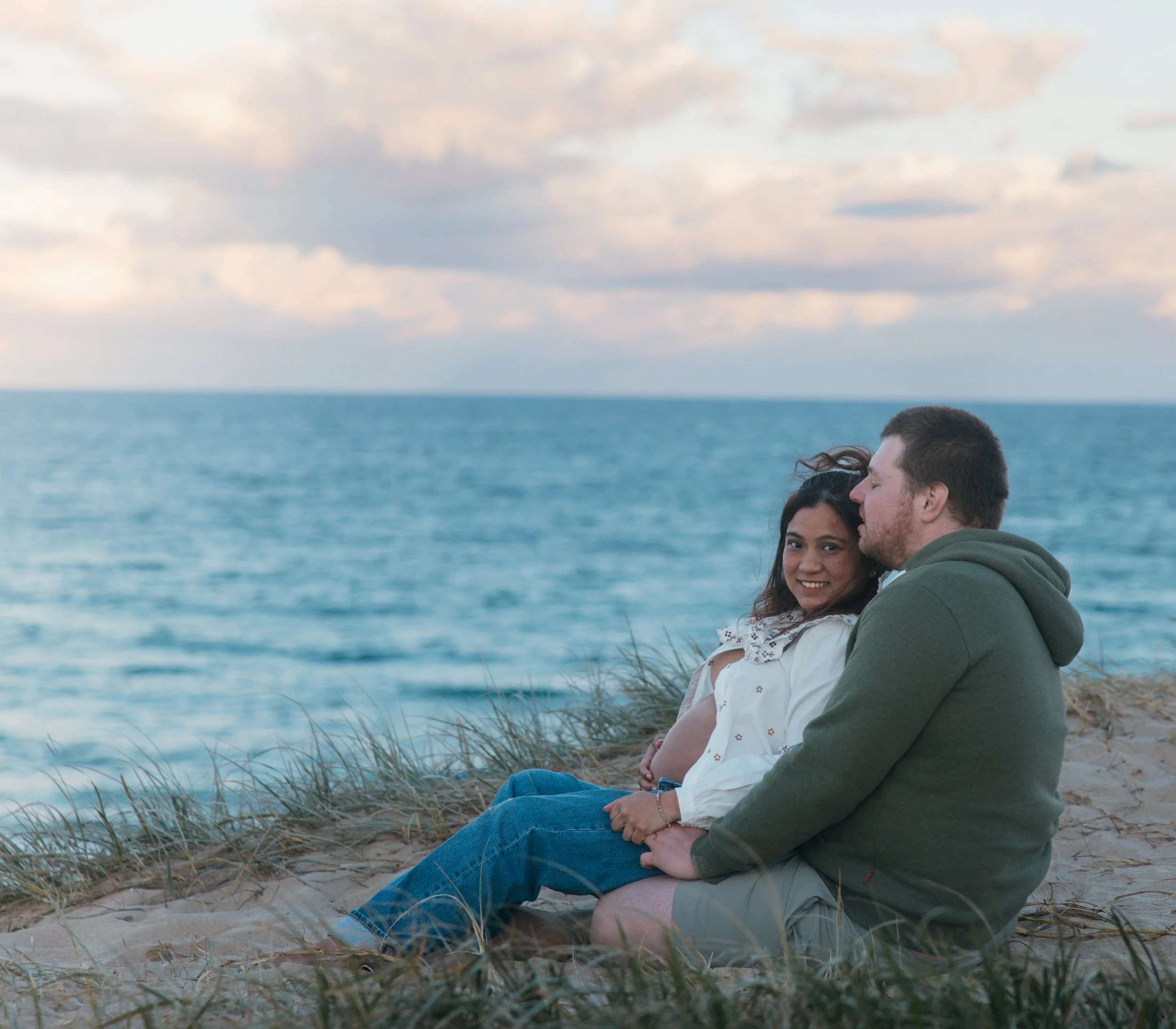 A couple sitting on the beach, with a woman smiling and a man whispering into her ear, near the ocean with cloudy sky in the background.