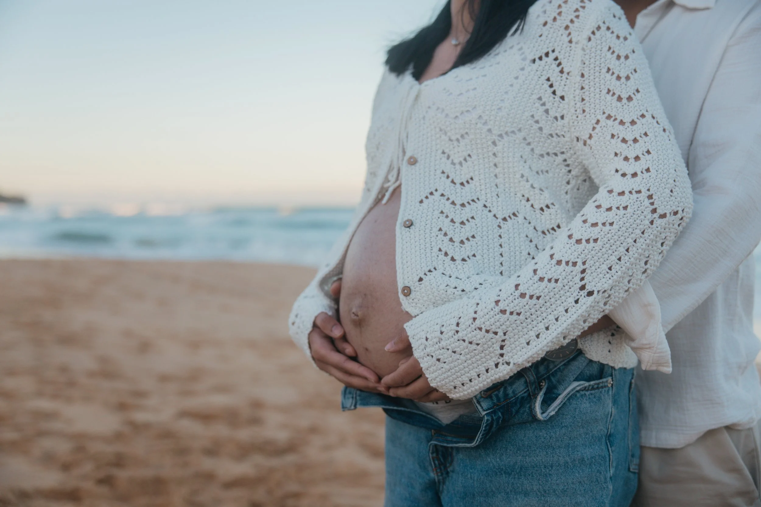 Pregnant woman in a white crochet cardigan holding her belly on a beach with waves in the background.