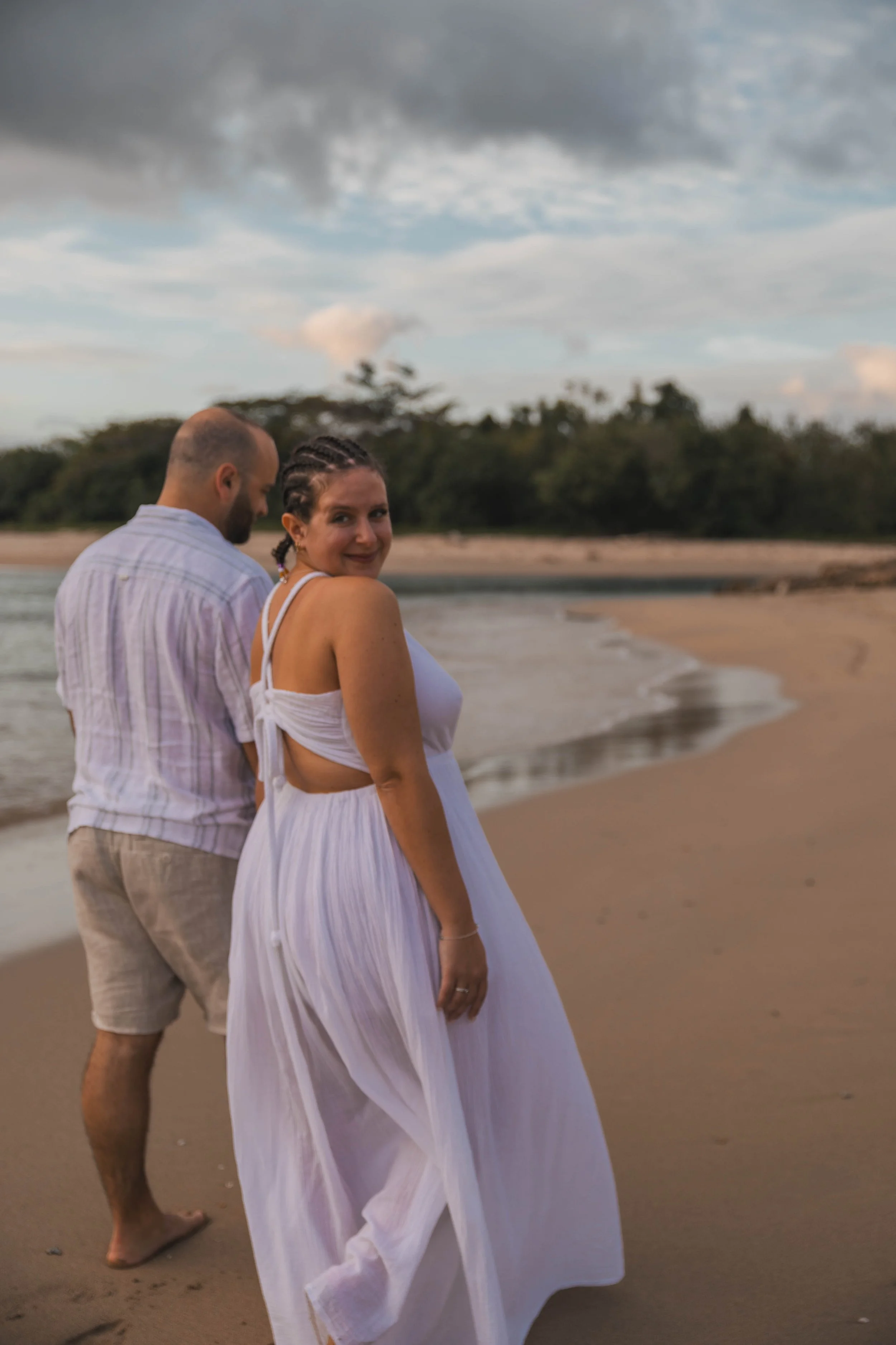 A woman in a white dress smiling at the camera on a beach with a man behind her, with the shoreline, trees, and cloudy sky in the background.