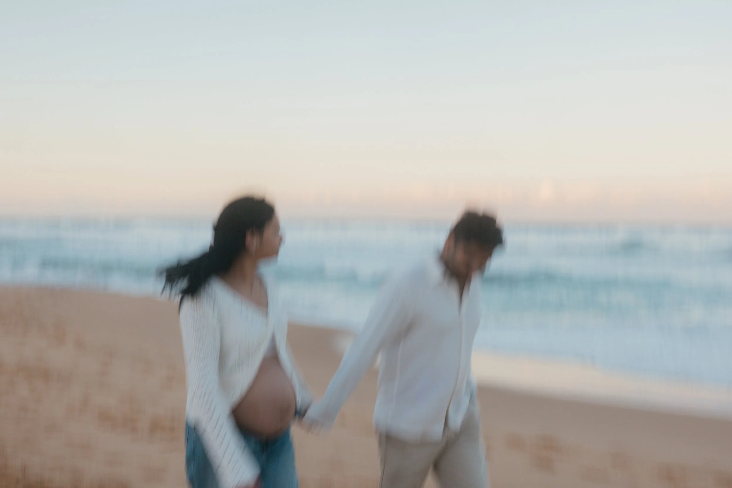 A blurry photo of a pregnant woman and her partner holding hands while walking on the beach with ocean waves in the background during sunset.