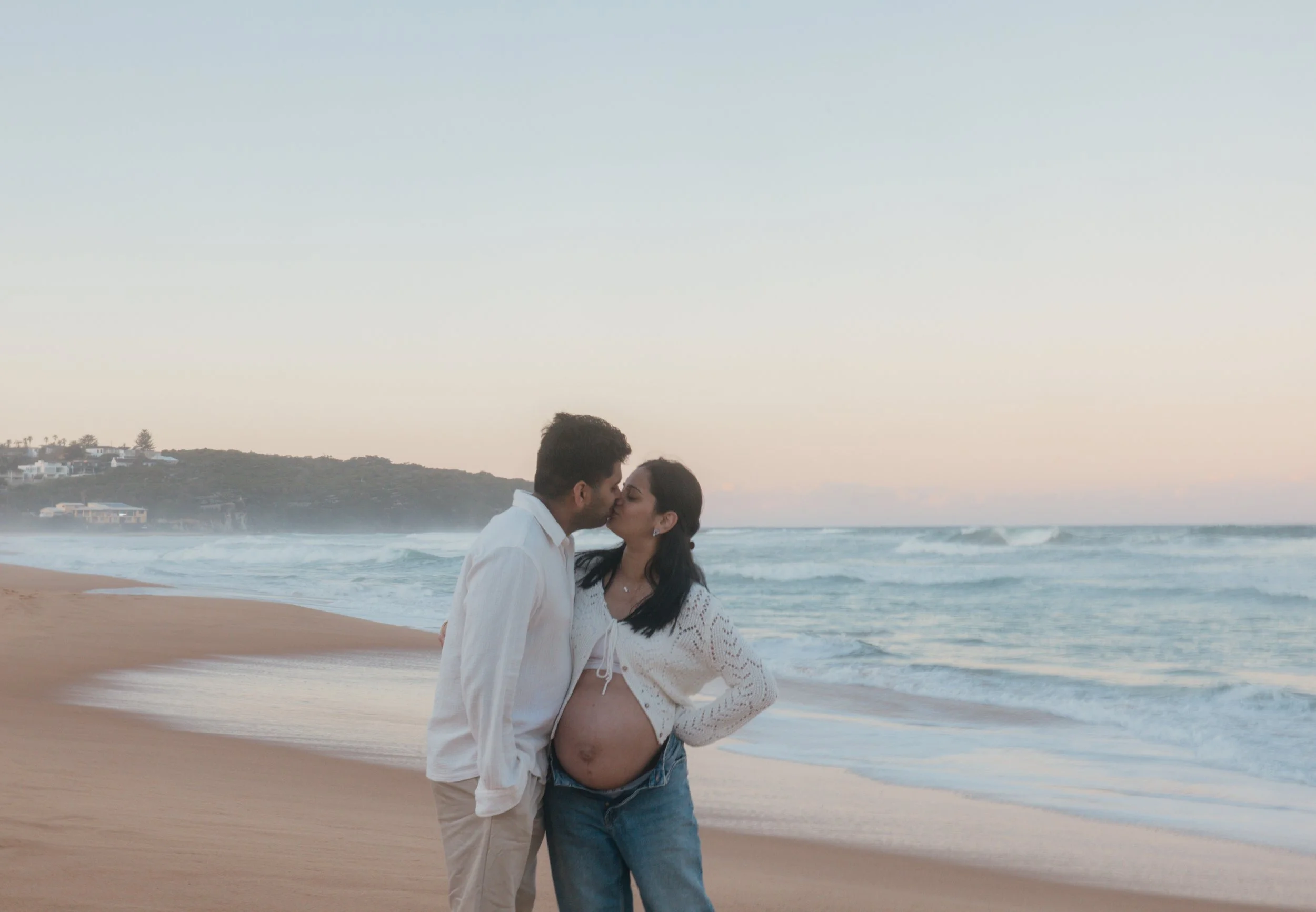 Couple kissing on the beach, woman with a pregnant belly, ocean waves in the background.