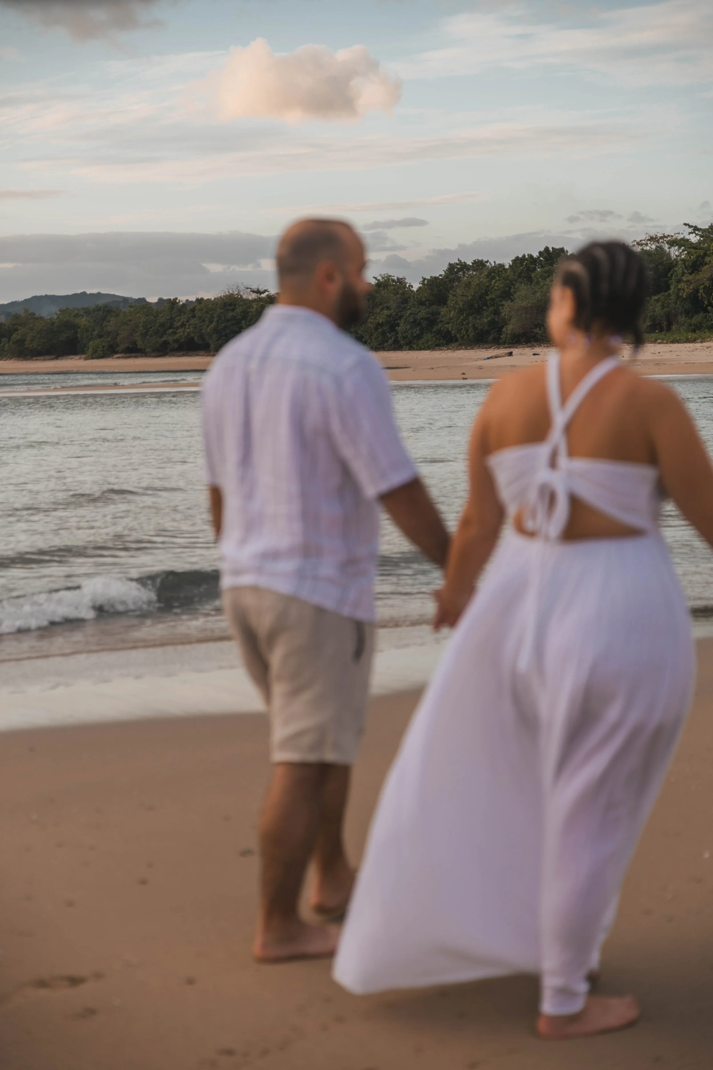 A man and woman holding hands while walking on a sandy beach near the ocean during sunset, with a cloudy sky and distant tree-covered hills.