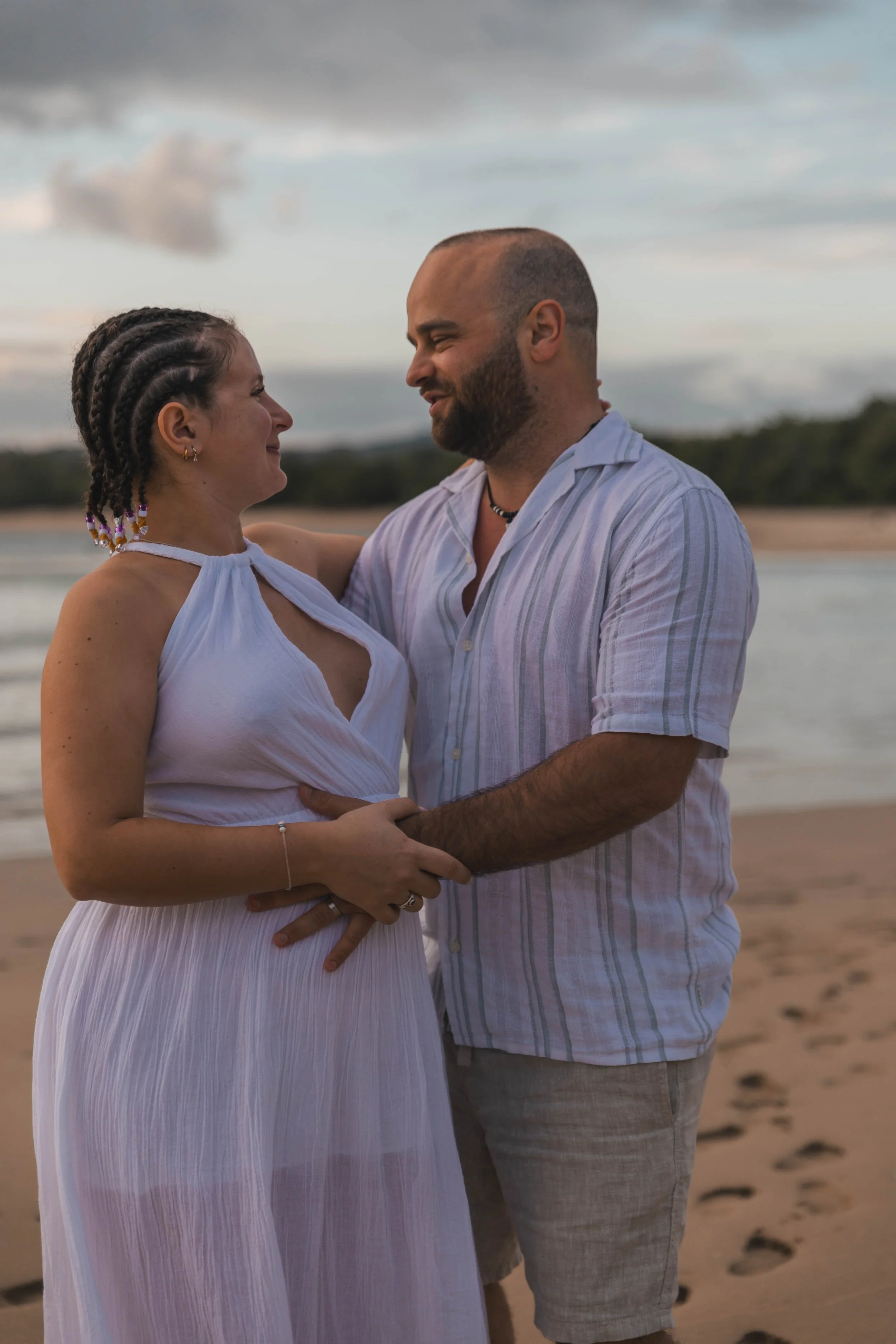 A couple standing close together on a beach, smiling and gazing into each other's eyes during sunset.