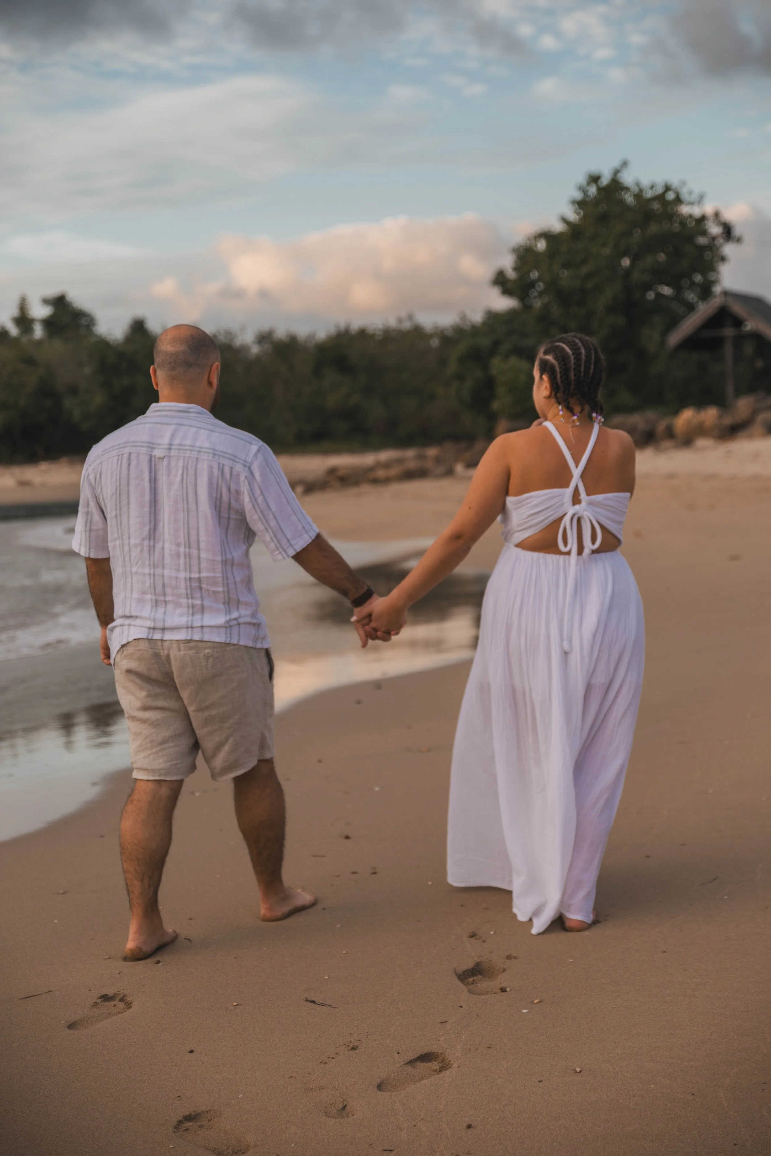 A couple walking hand in hand on a sandy beach during sunset, leaving footprints in the sand with trees and a small house in the background.