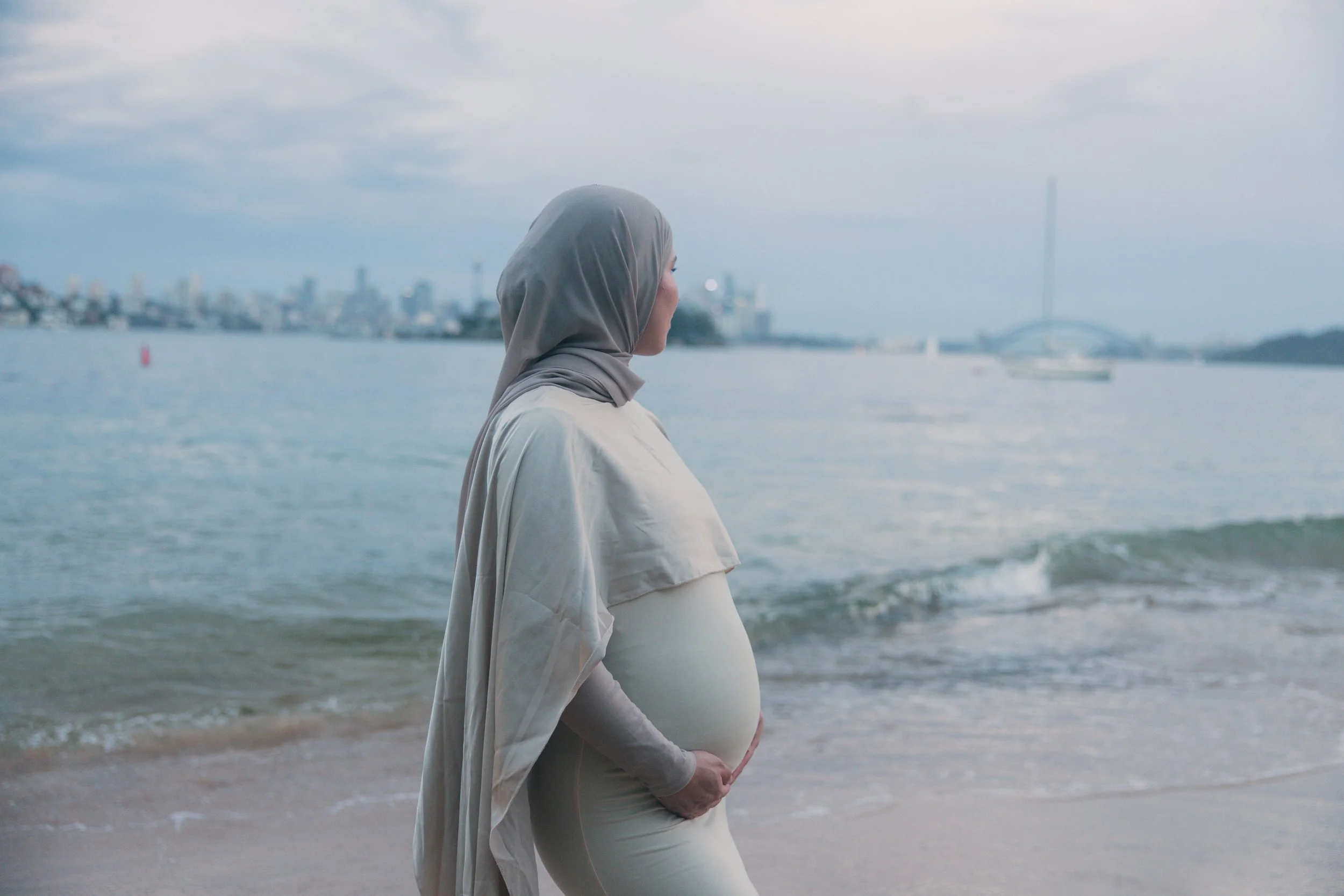 Pregnant woman wearing a hijab standing on the beach, looking at the water with a city skyline and bridge in the background.