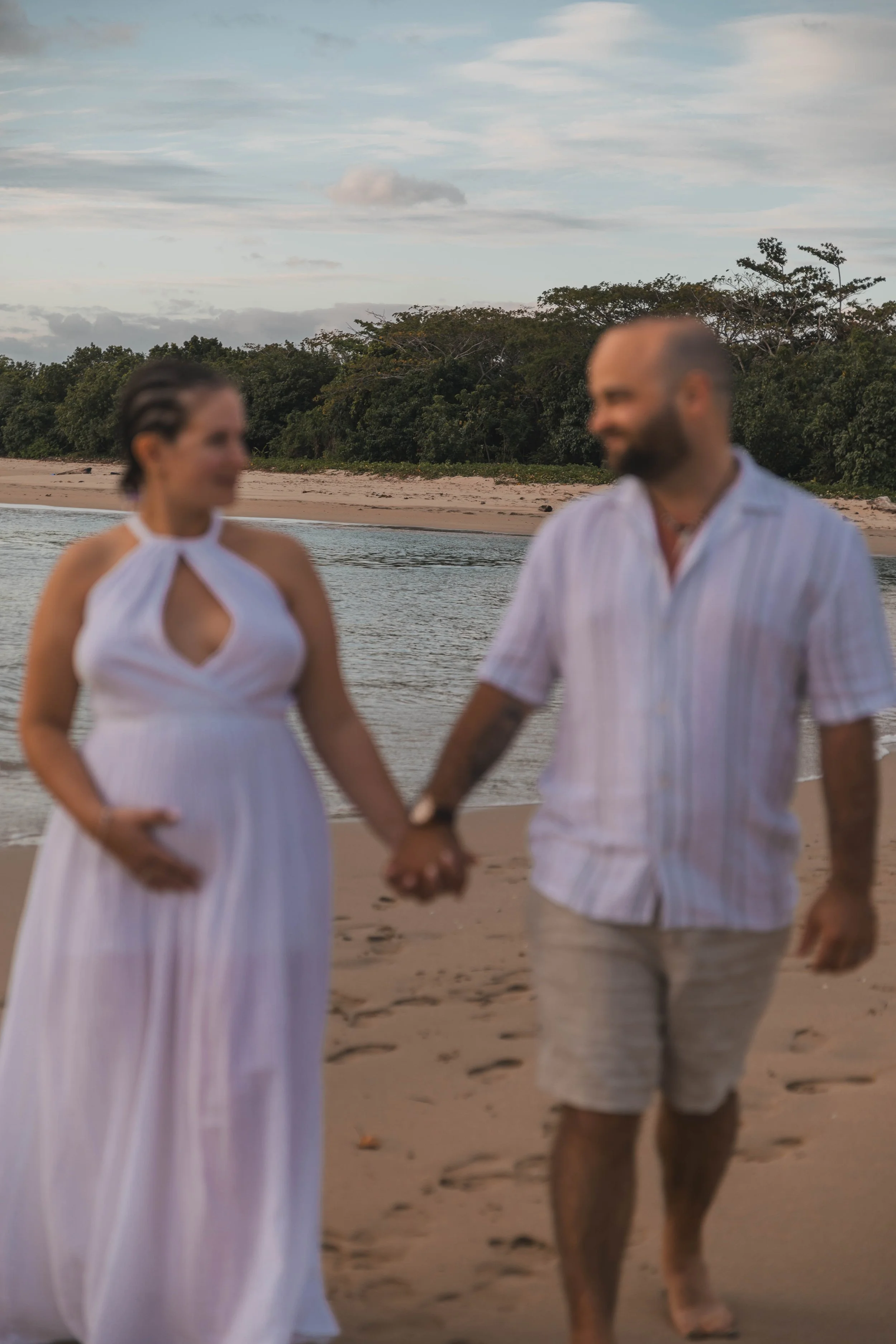 A couple walking hand in hand on the beach during sunset, with trees and the ocean in the background.