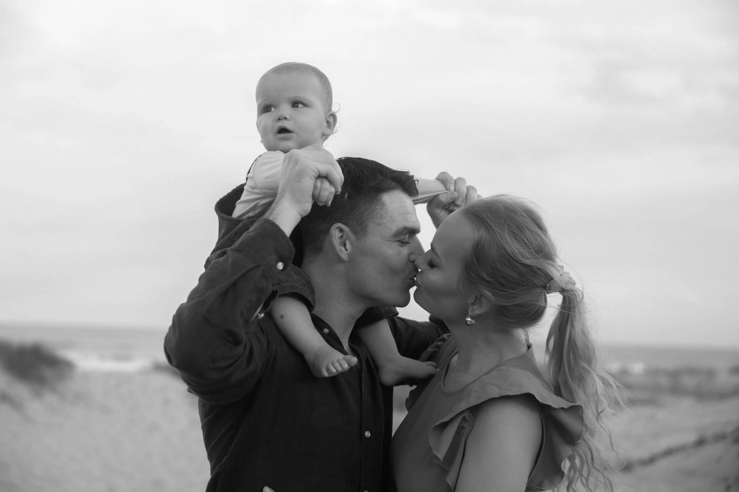 A couple kissing with a young child sitting on the man's shoulders at the beach.