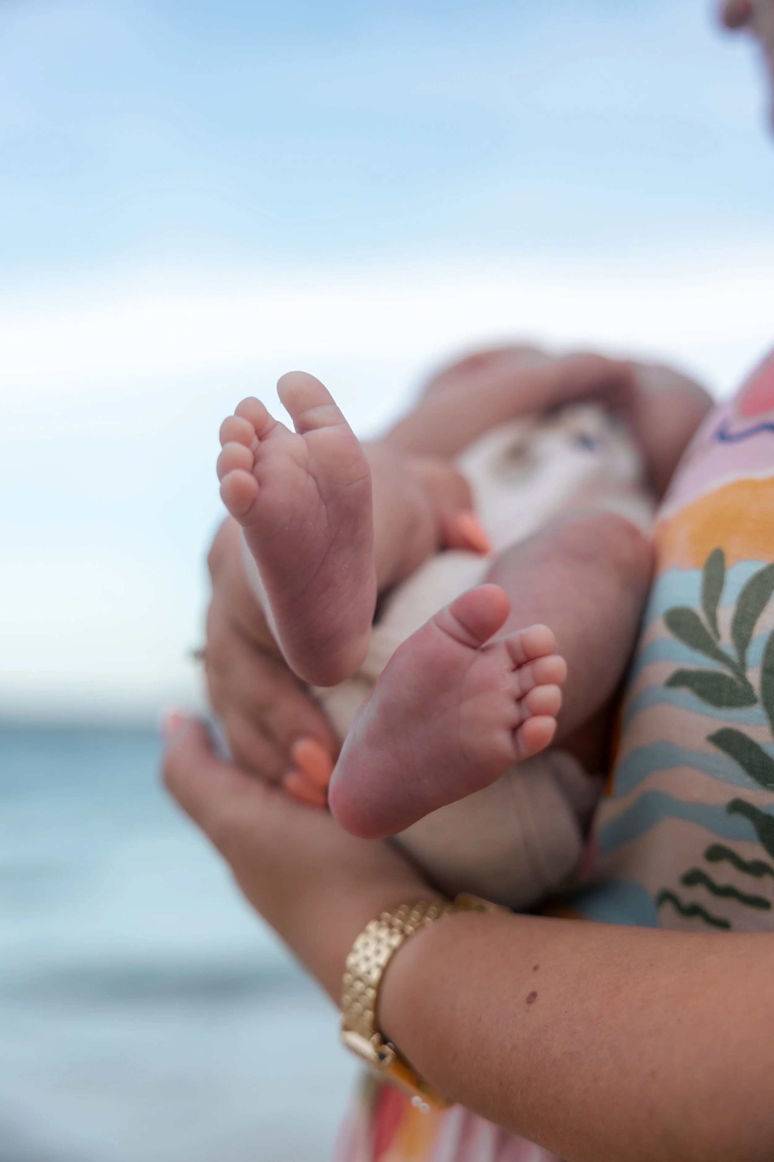 Close-up of a woman holding a newborn baby against a background of a blue sky and water, focusing on the baby's tiny feet and hands.