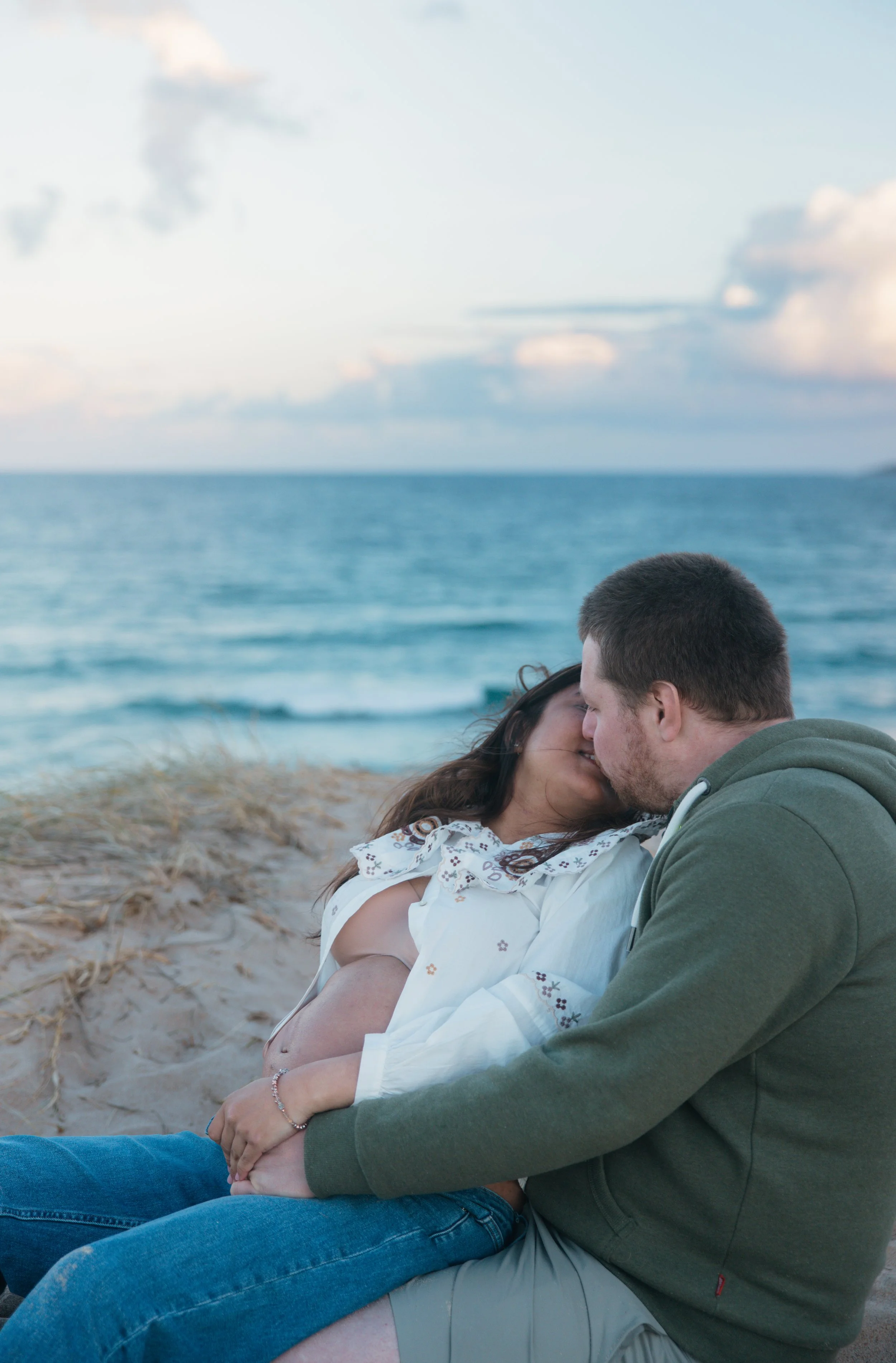 A couple sitting on the beach, kissing, with the ocean and sky in the background.
