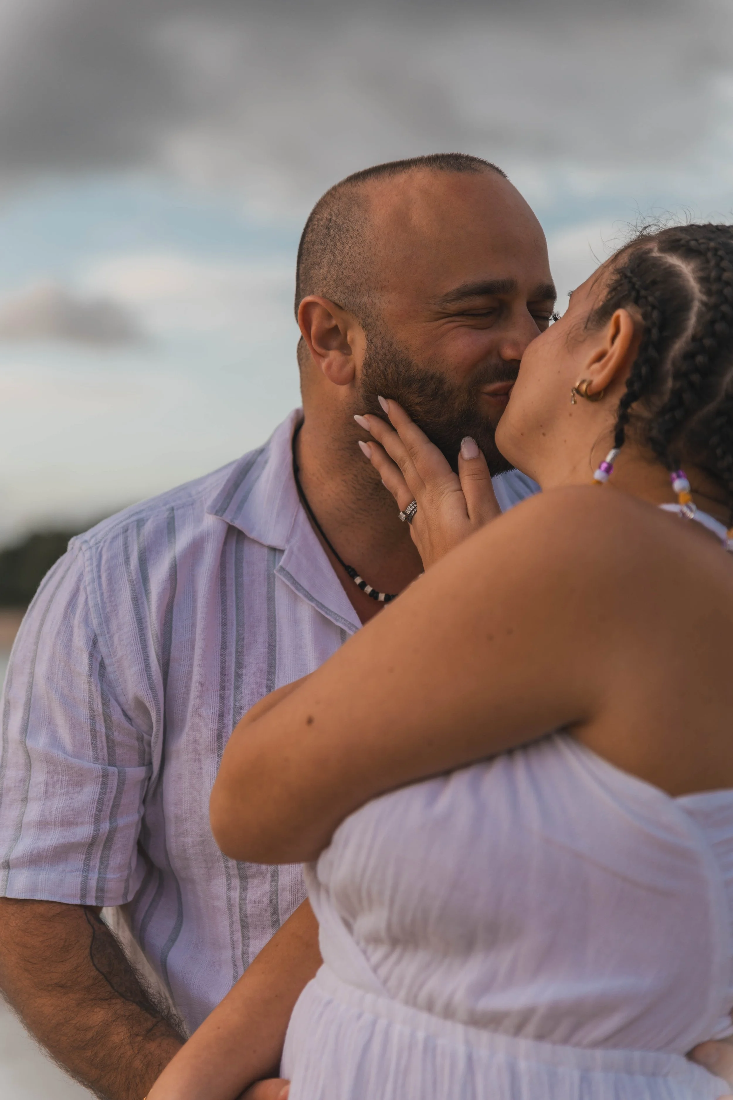 A couple sharing a kiss outdoors, with the woman touching the man's face, both with joyful expressions. The man has a beard and is wearing a striped shirt, while the woman has braids and colorful jewelry.