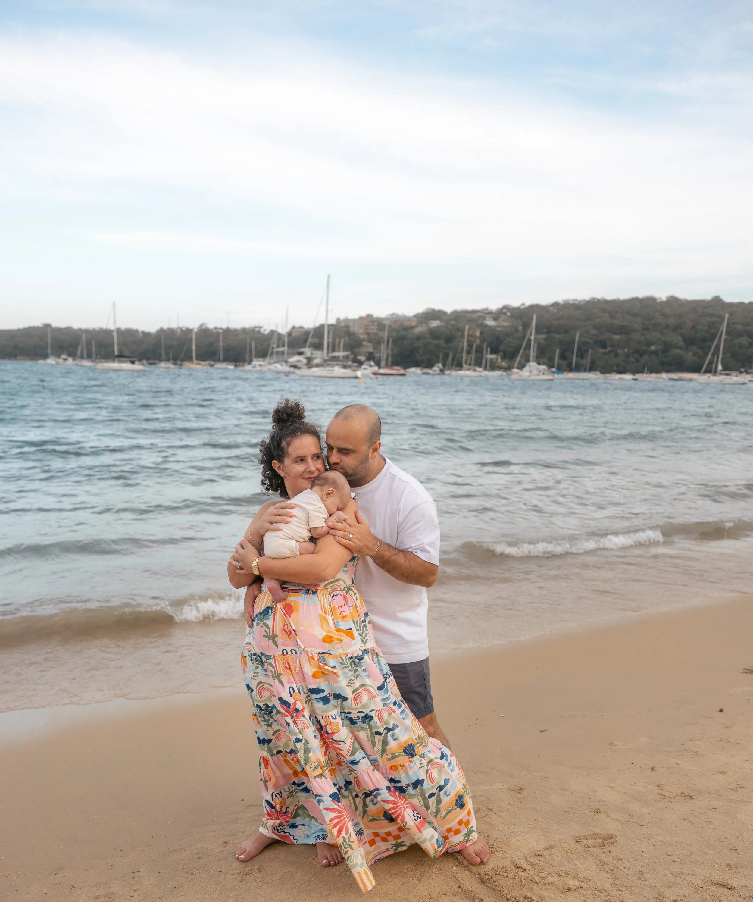 A family of three, consisting of a woman, a man, and a baby, stands on a sandy beach near the water, with boats and a hill in the background, during daytime.