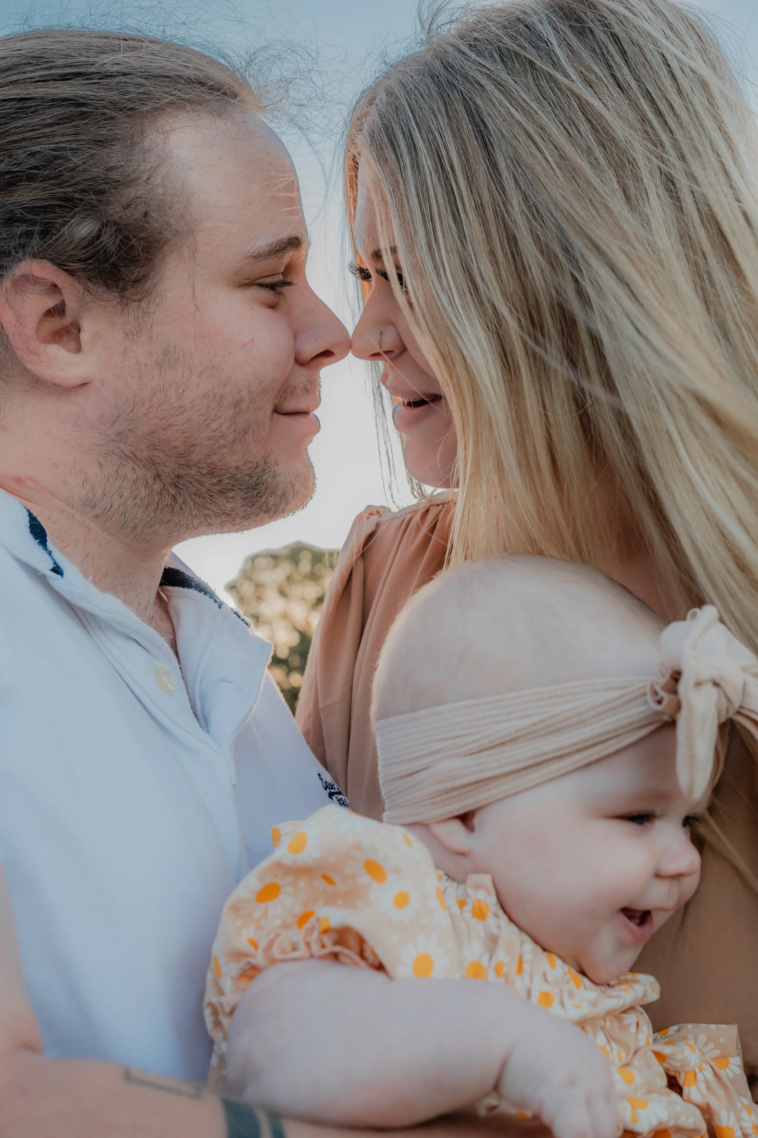 A young couple with a baby baby embracing outdoors as they touch foreheads, smiling, with a blurred background of trees and sky.