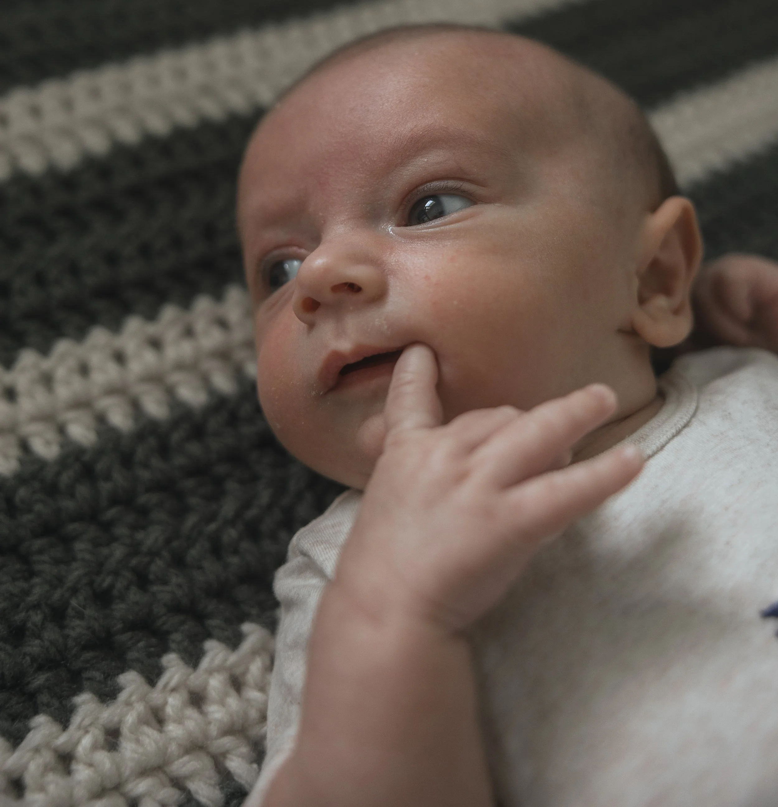 Close-up of a baby lying on a textured blanket, with a finger near their mouth, looking off to the side.