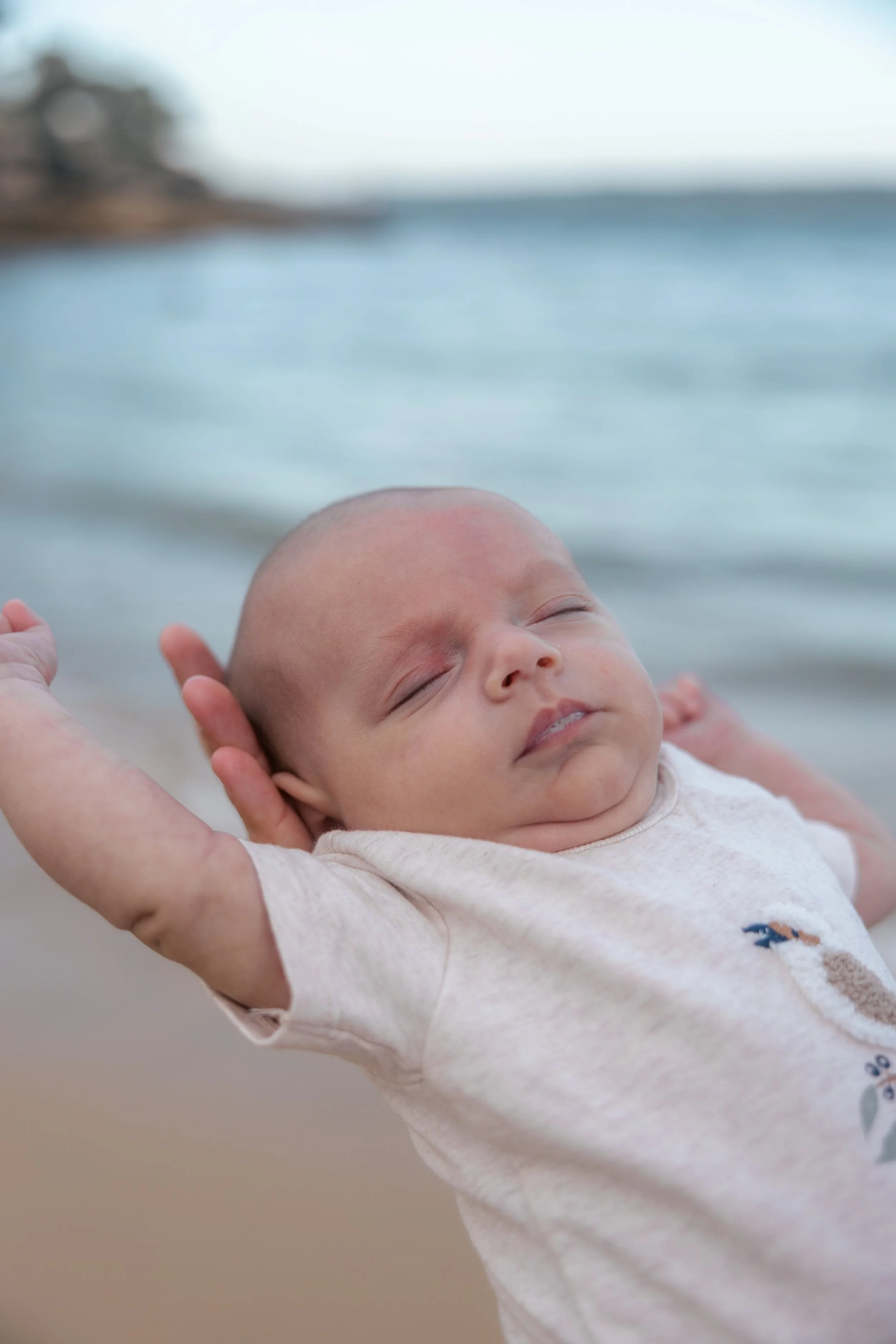 A sleeping baby with arms stretched back, lying on someone at the beach with the ocean in the background.