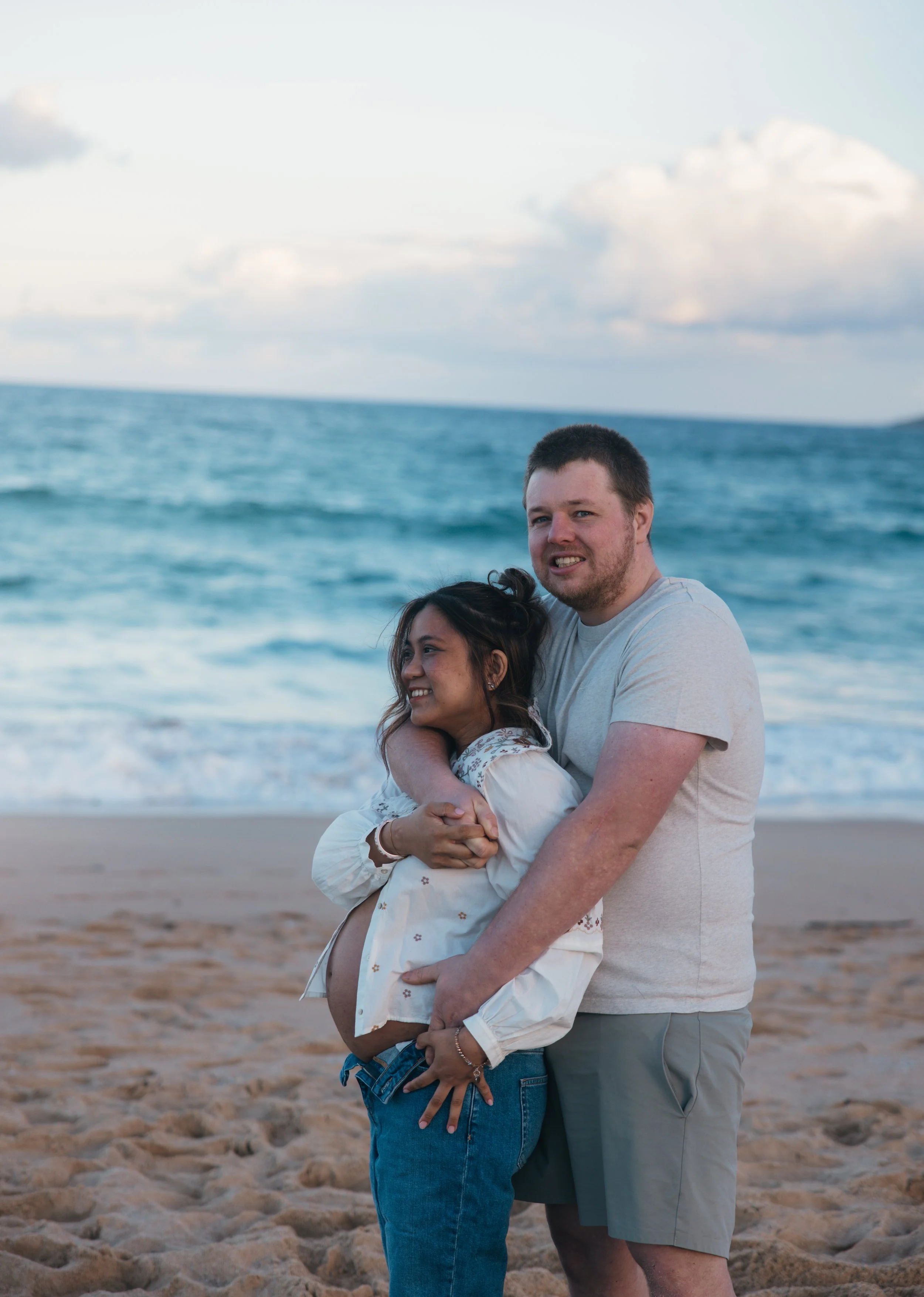 A couple hugging on the beach with the ocean in the background, both smiling.
