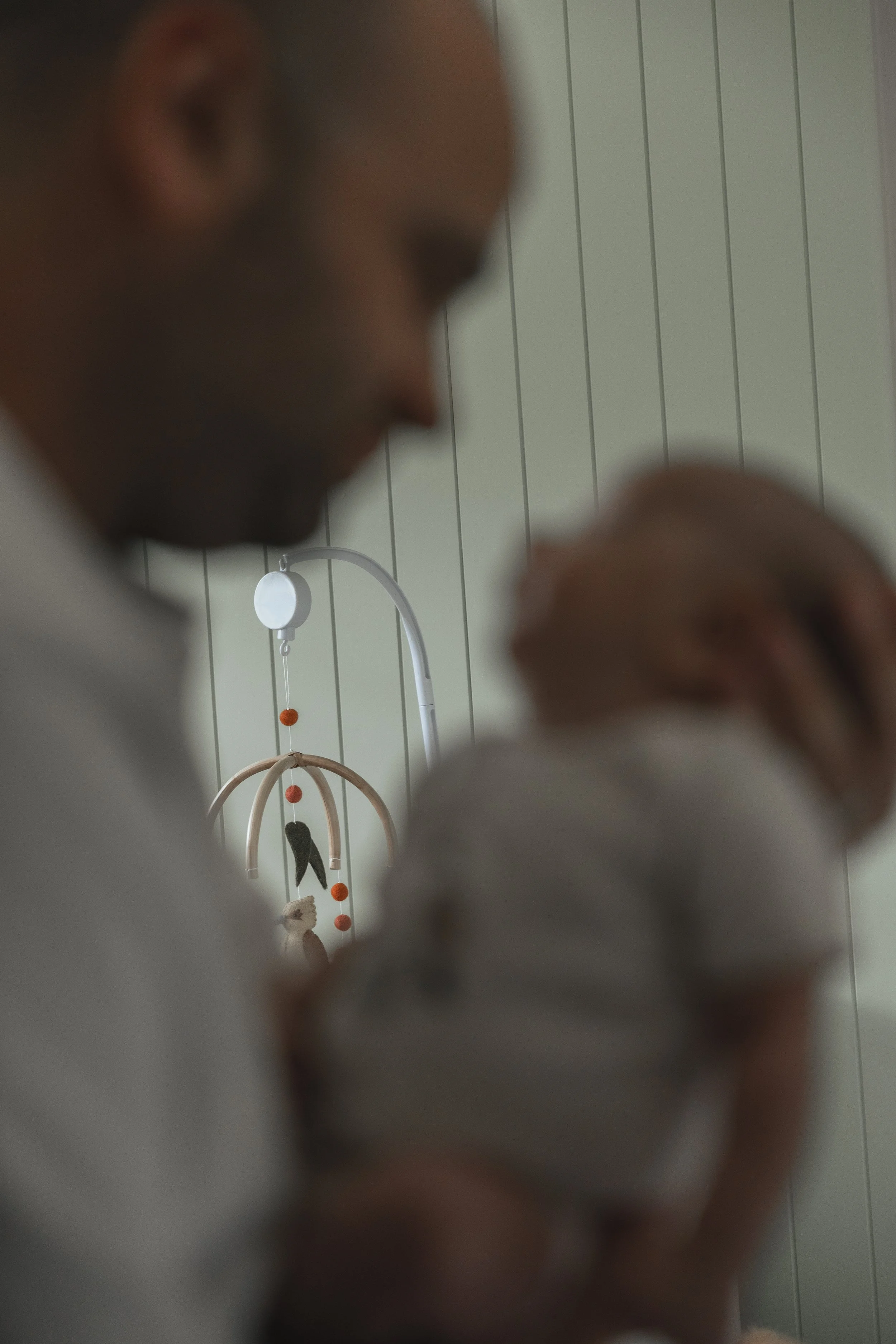 A person holding a newborn baby in a room with a white wall. A mobile with orange and black decorations hangs in the background.