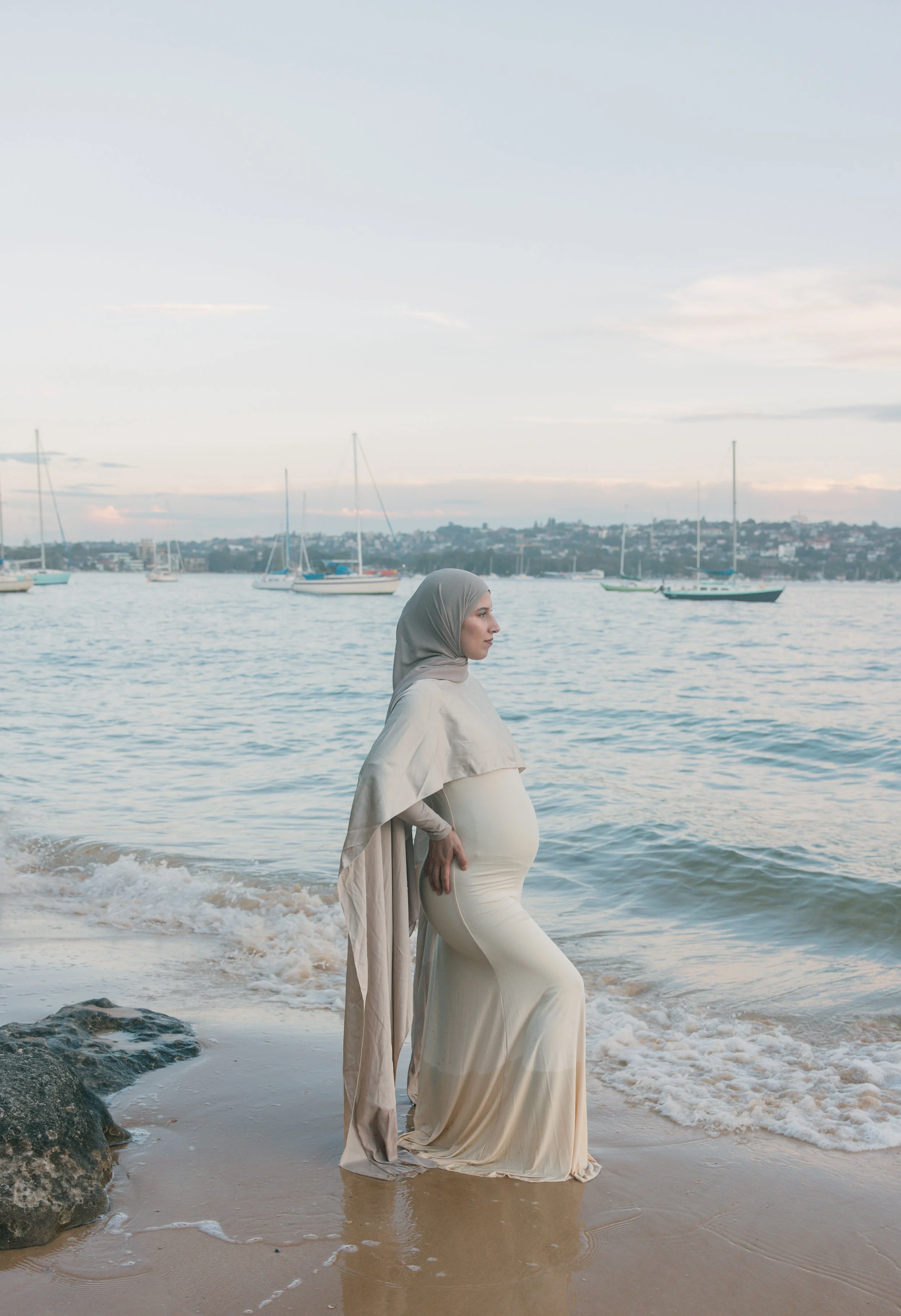 A woman wearing a hijab and flowing dress standing on a beach with her hands on her pregnant belly, facing the water with boats in the background.