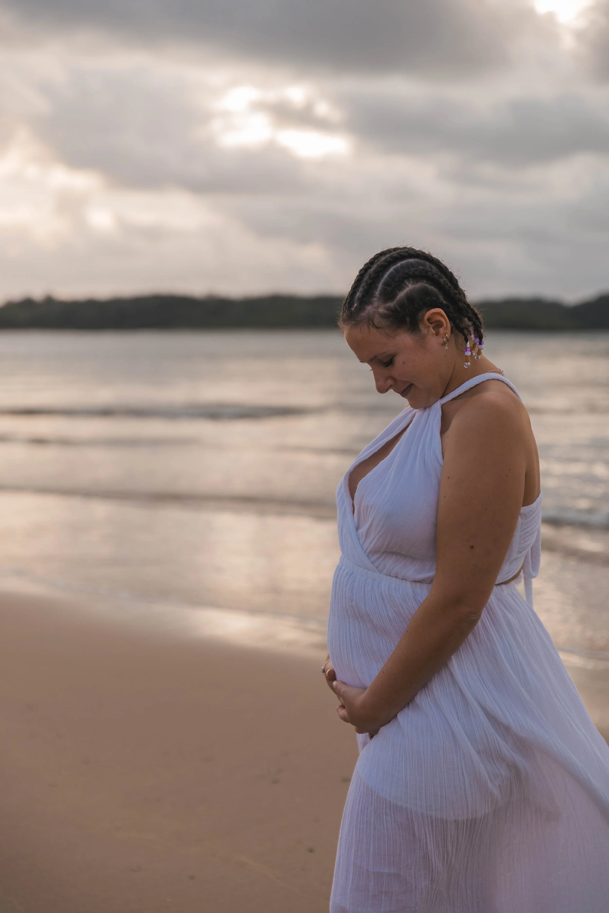 Pregnant woman standing on a beach in a white dress, looking down, during cloudy weather.