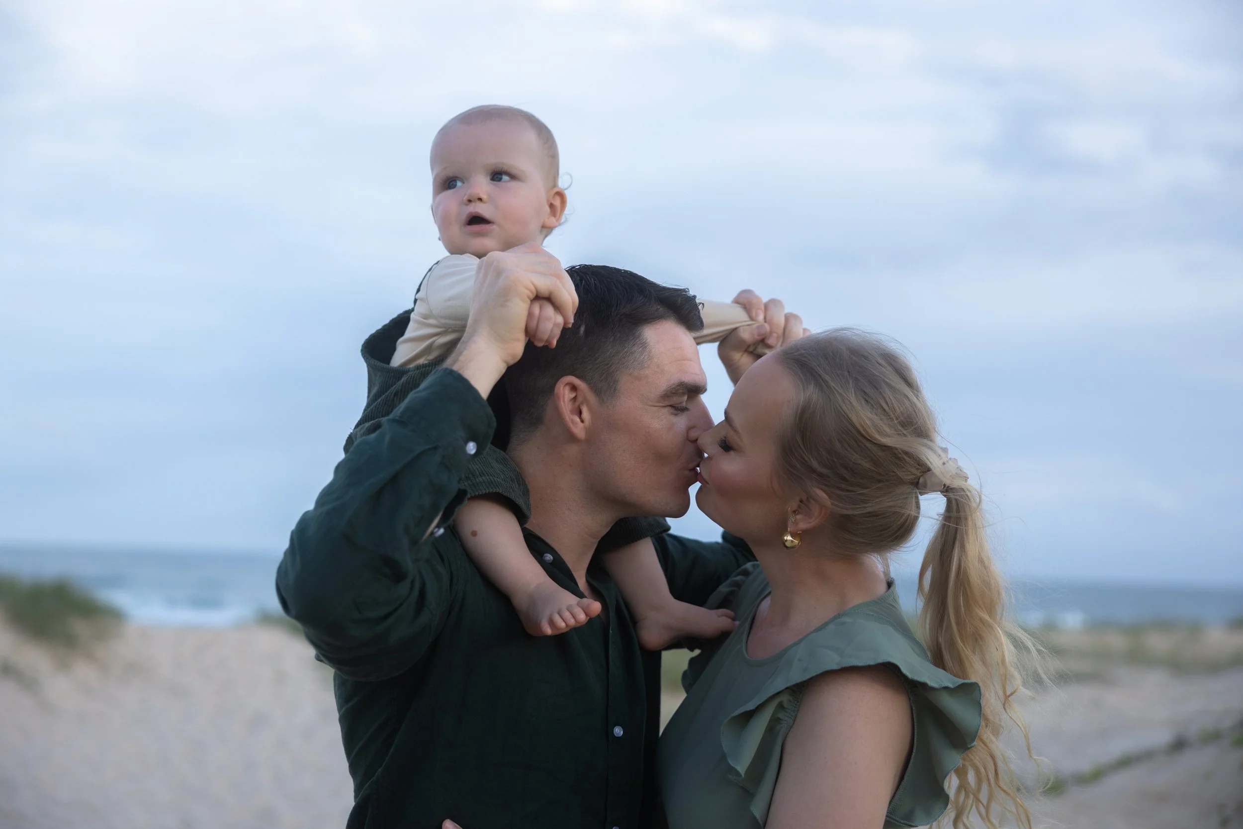 A couple kissing on a beach with a man carrying a baby on his shoulders, and the ocean in the background.