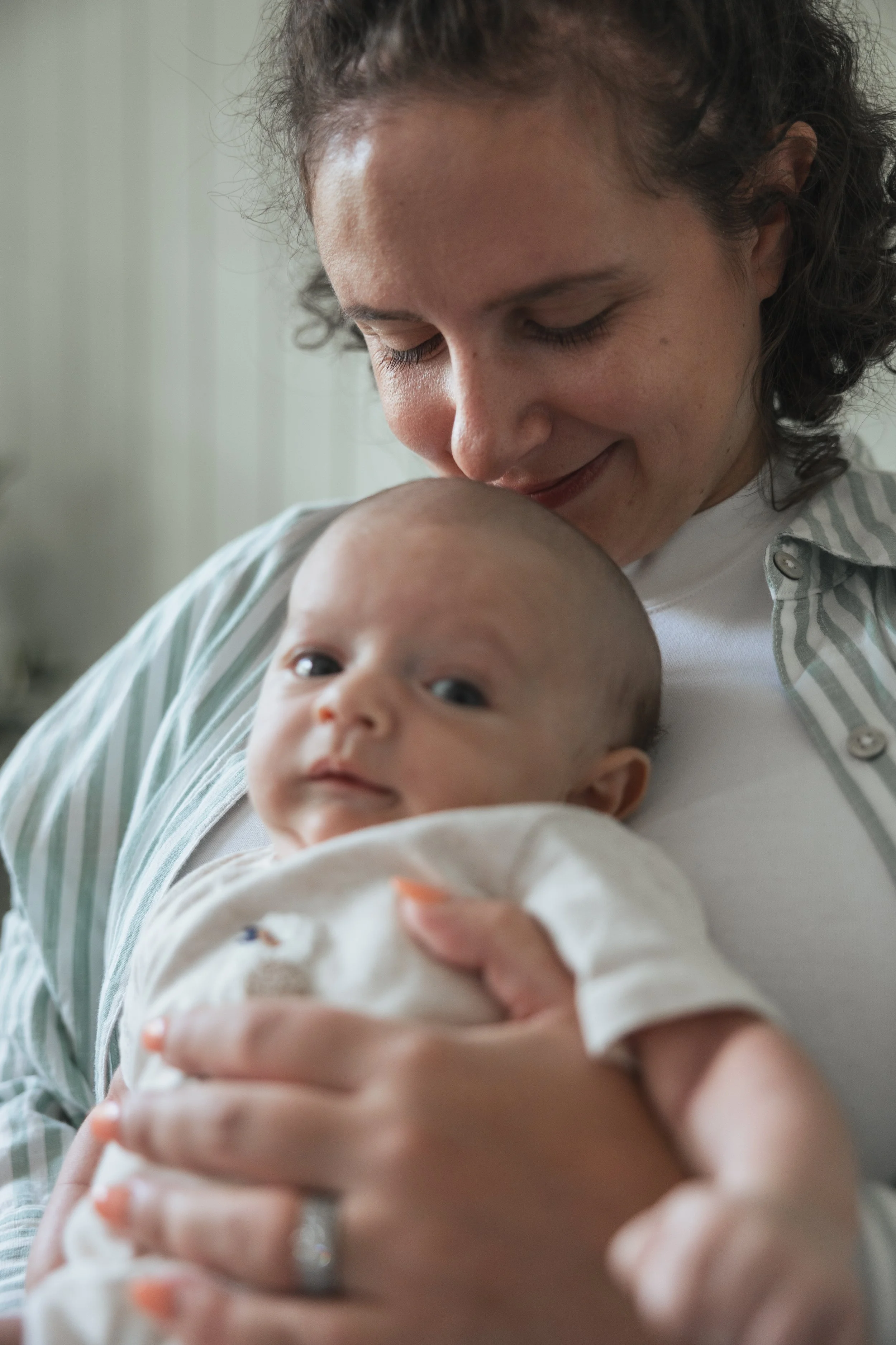 A woman holding a baby close to her chest, both smiling softly. The woman is gently looking down at the baby, who is looking at the camera.