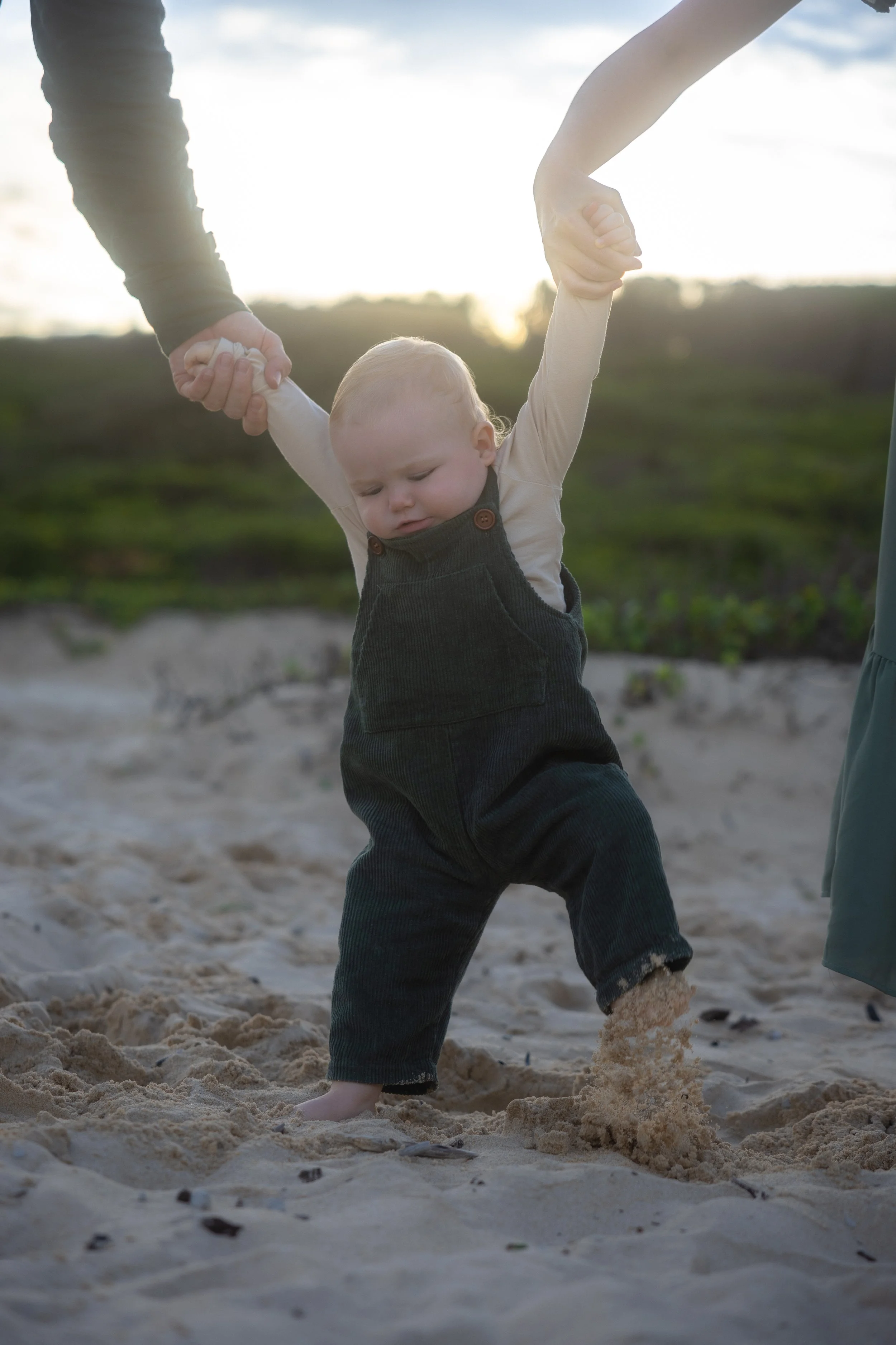 A toddler walking on sandy beach while holding two adults' hands, with the sunset in the background.