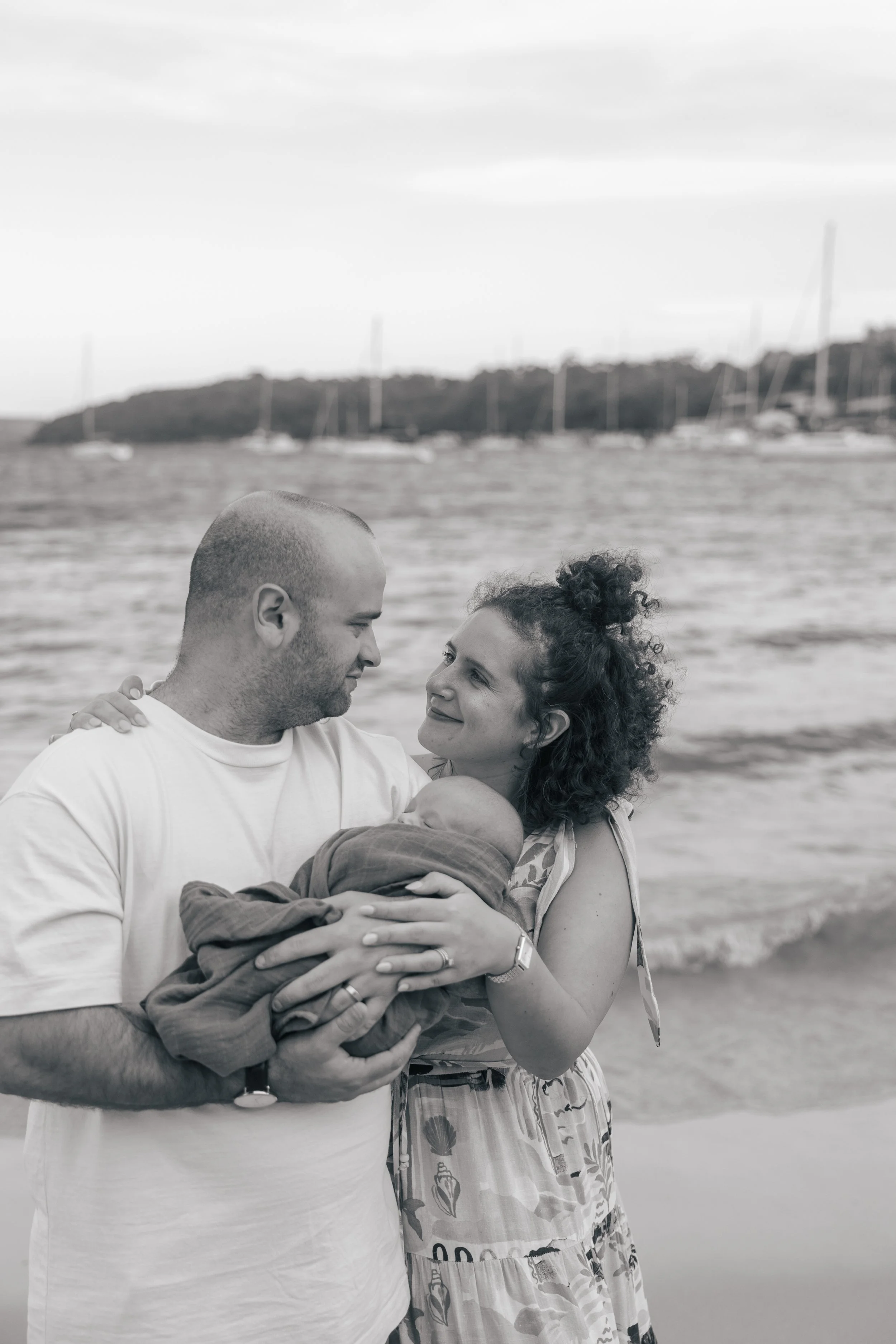 A black-and-white photo of a family on the beach. A man and woman are holding a baby wrapped in a blanket, looking at each other affectionately with the ocean and boats in the background.
