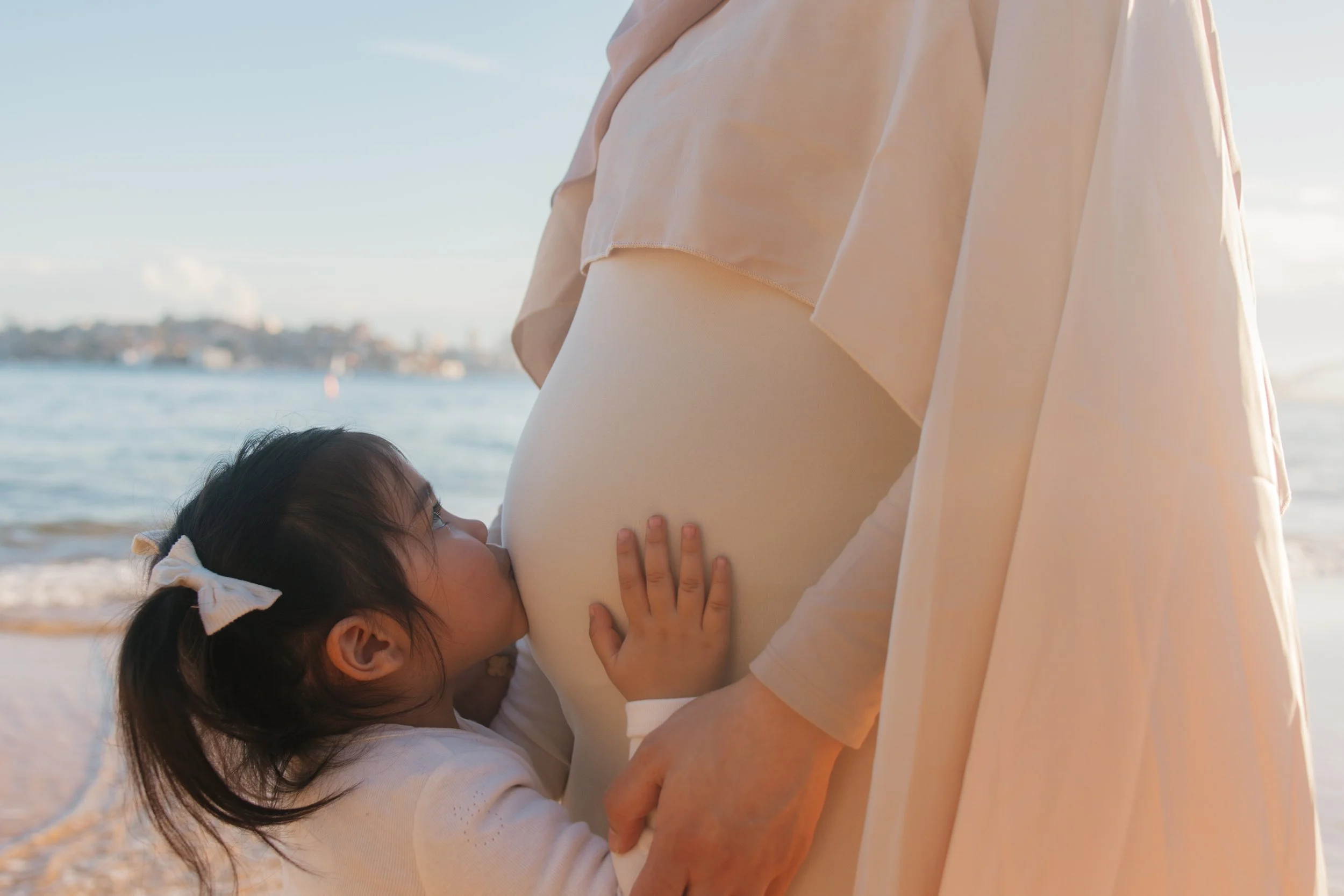 A young girl kisses a pregnant woman's belly on the beach during sunset.