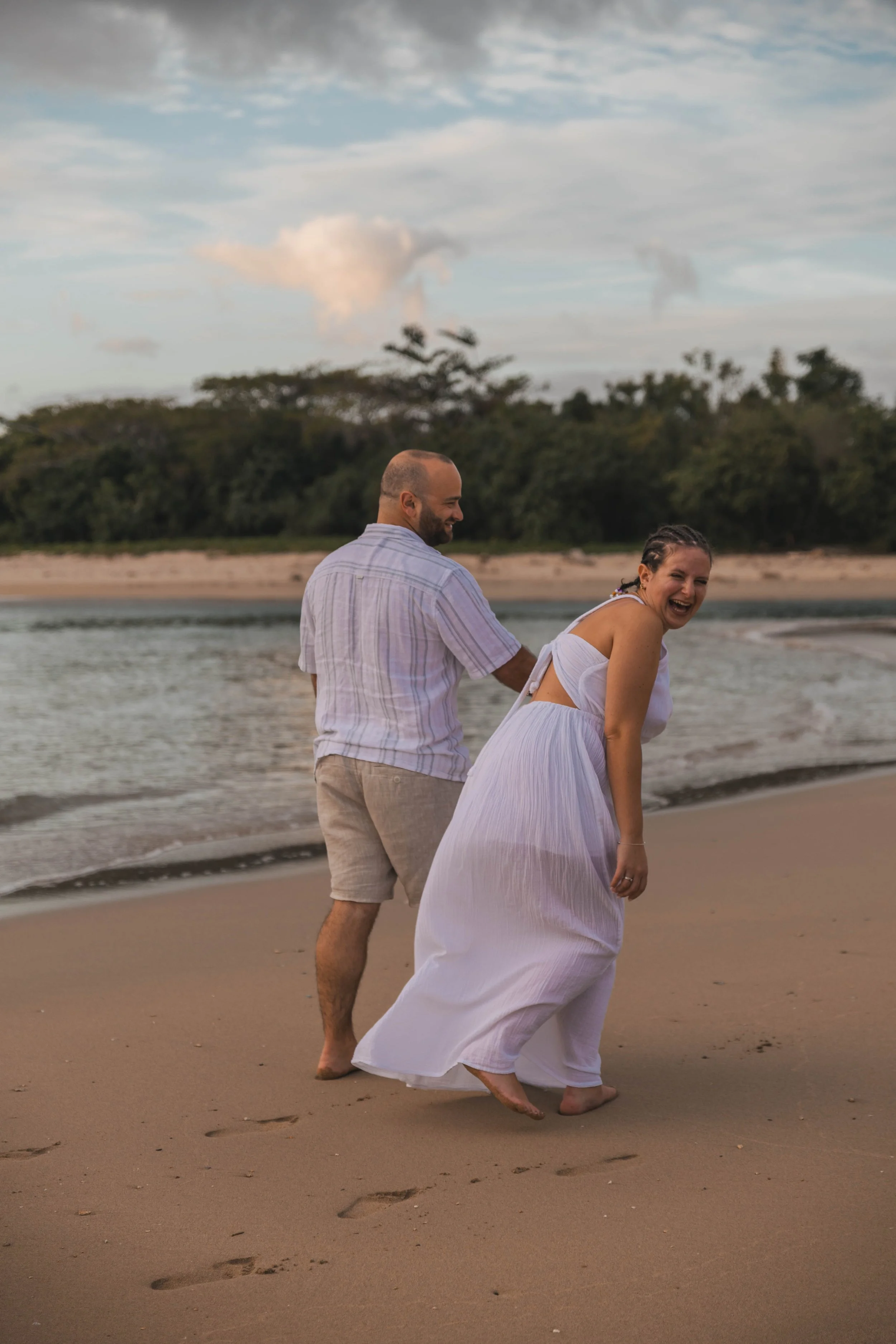A man and a woman laughing and playing in the sand on a beach, with the ocean and trees in the background during sunset.